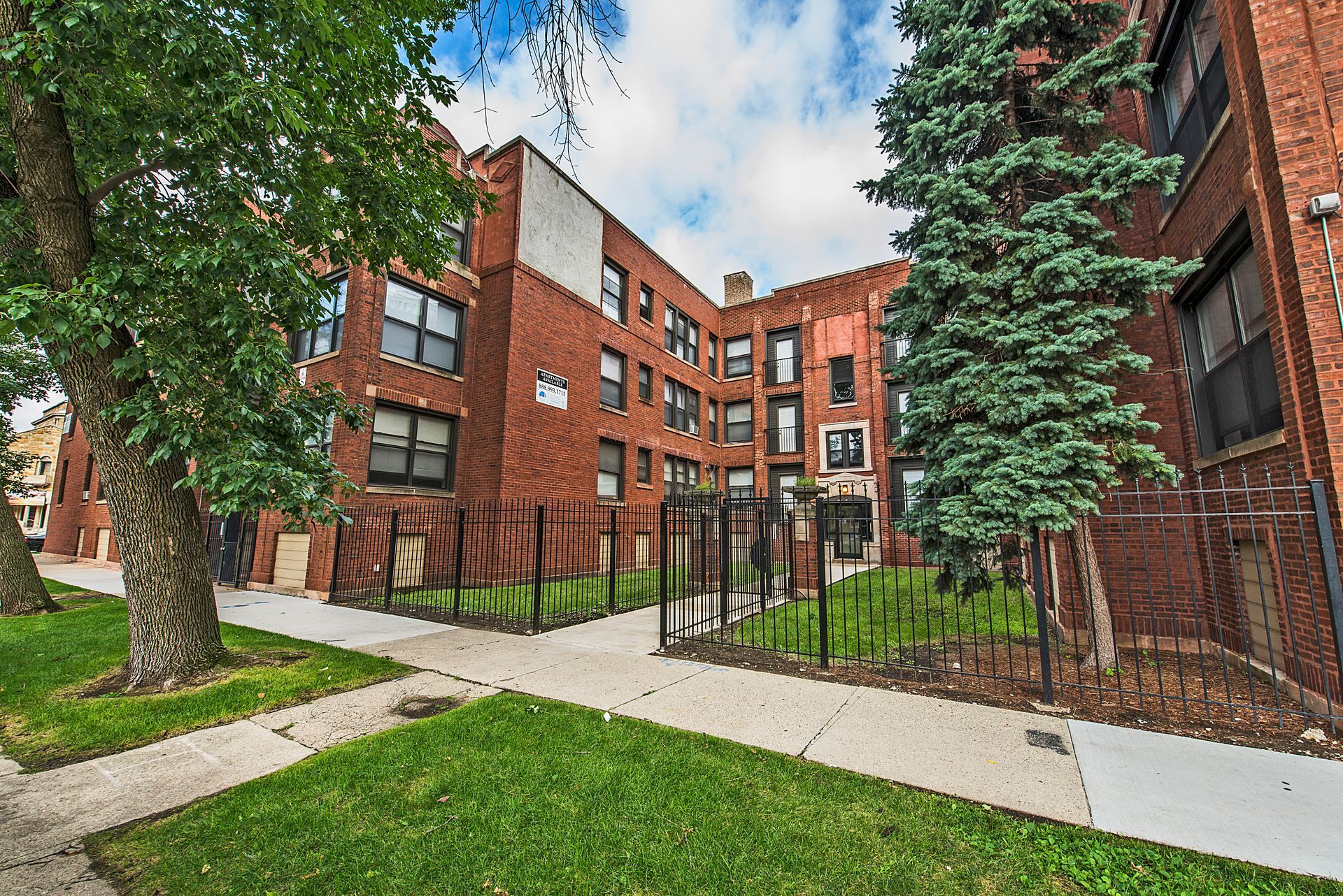 Brick apartment building with green lawn, trees, and fenced-in courtyard on a sunny day.