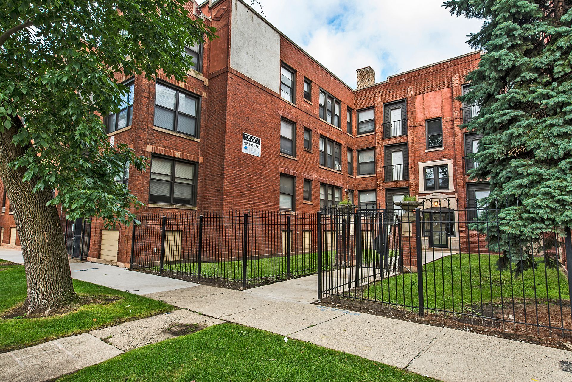 Red brick apartment building with black fence, trees, and walkway on a sunny day.