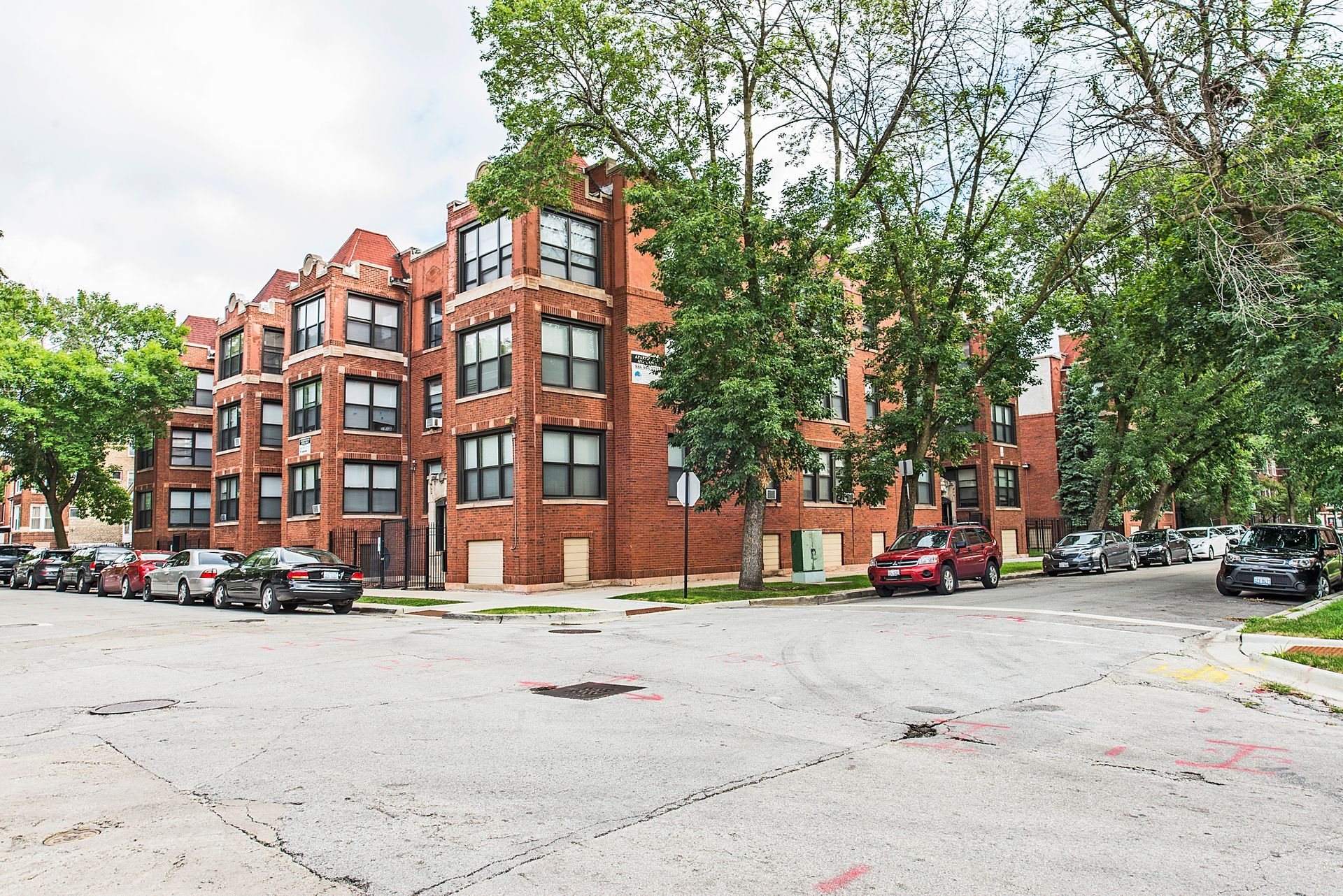 Brick apartment building on a corner lot, cars parked on the street, trees in front.