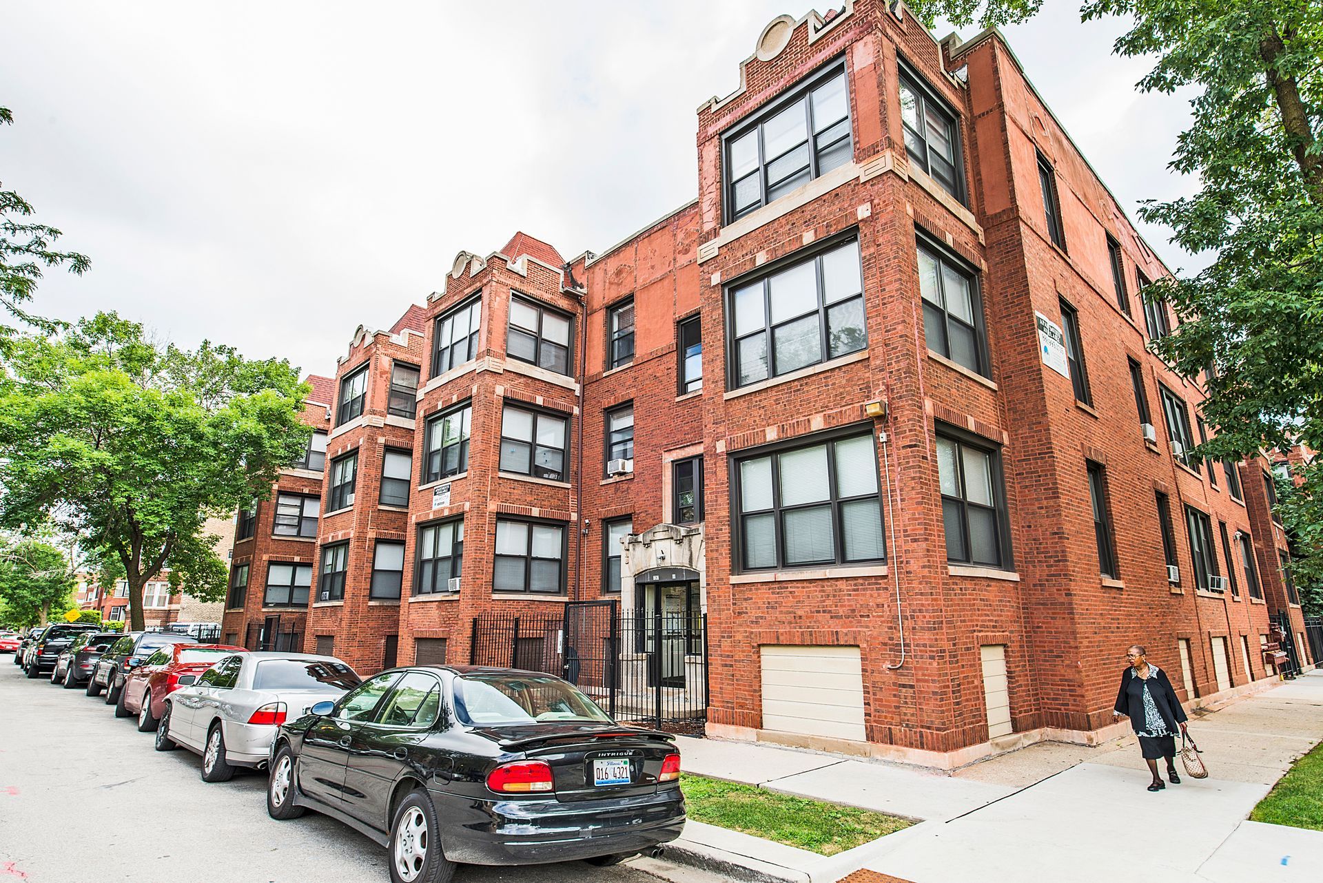 Red brick apartment building with parked cars on the street and a person walking on the sidewalk.