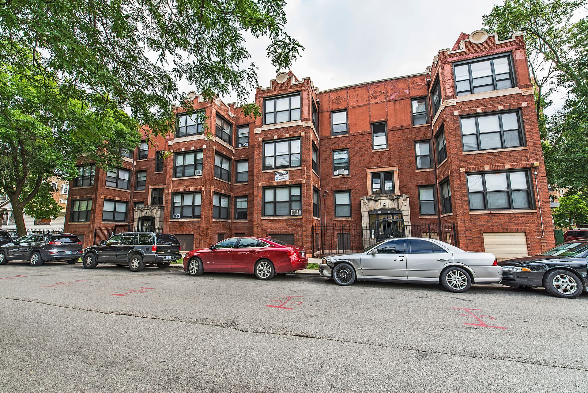 Red brick apartment building with cars parked on the street. Trees frame the edges.
