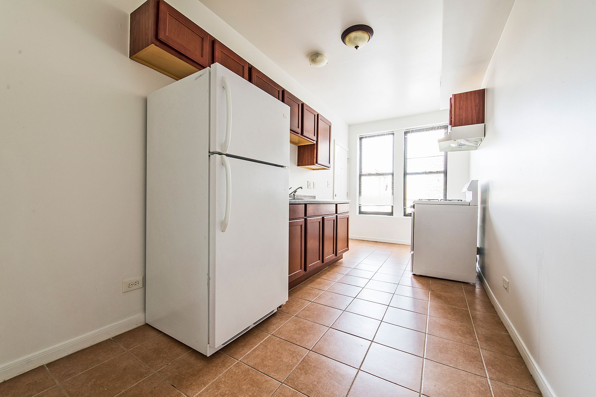 Empty kitchen with white appliances, brown cabinets, and tan tile floor.