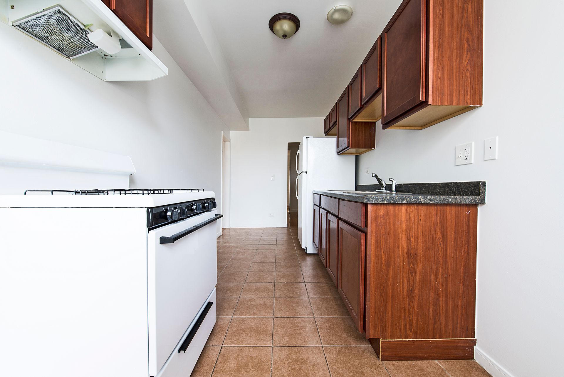 A narrow kitchen with white appliances, brown cabinets, and a tile floor.