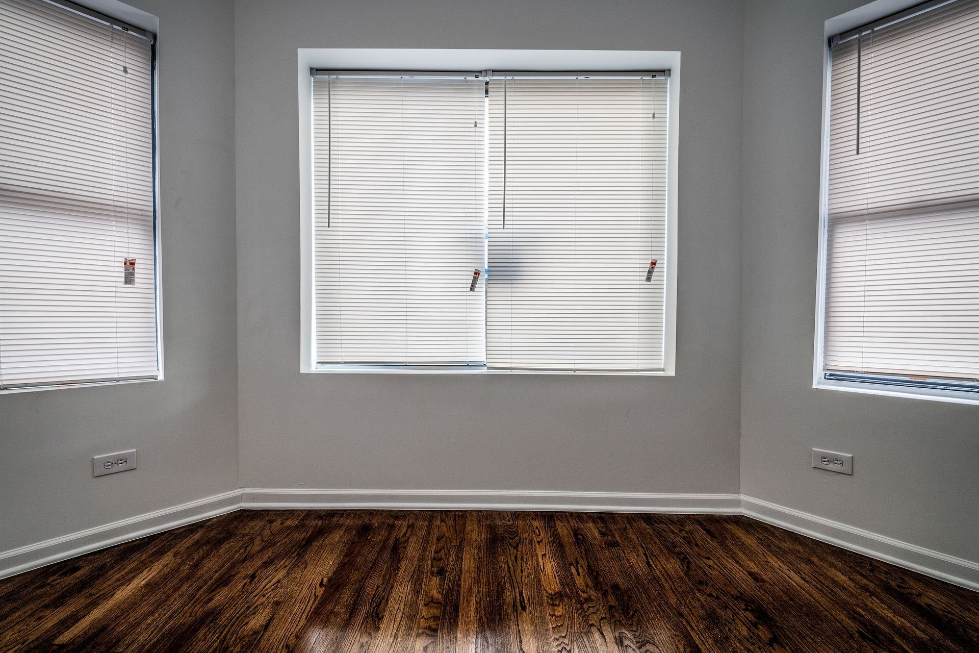 Empty room with three windows. White walls, wood floor, blinds drawn.
