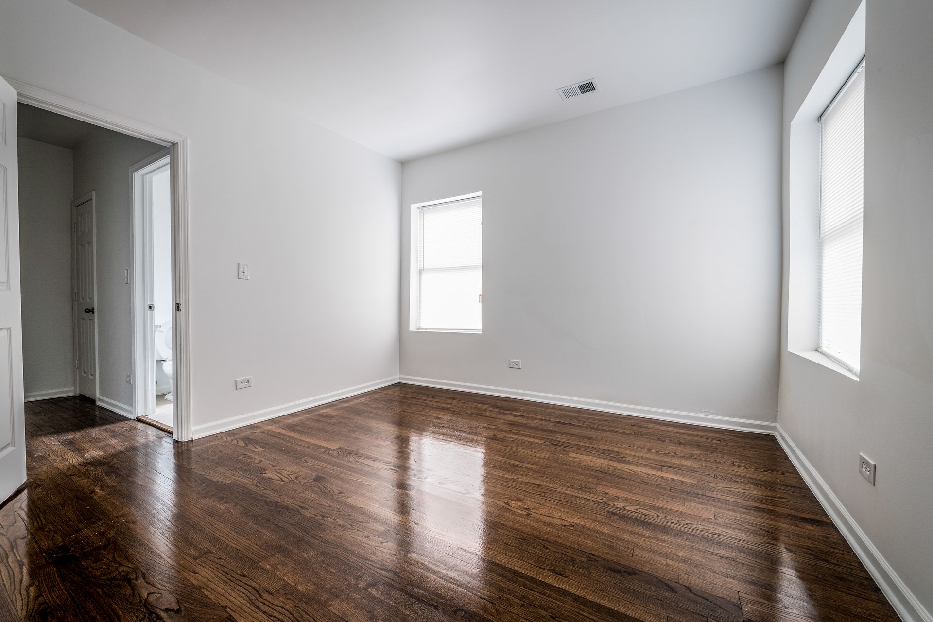 Empty room with hardwood floors, white walls, and two windows. A door is visible to the left.