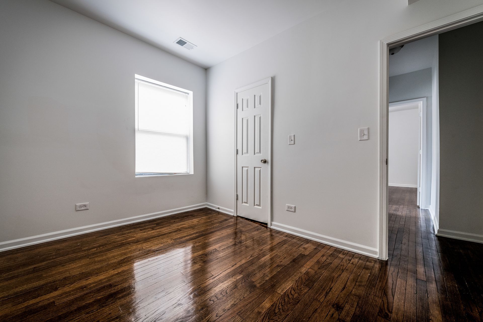 Empty room with hardwood floors, a white door, and a window; another doorway is visible.