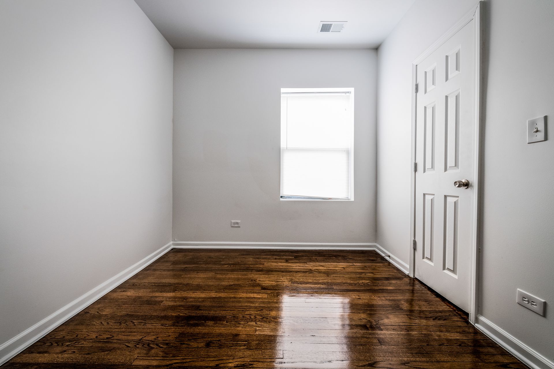 Empty room with hardwood floors, a closed white door, and a window.