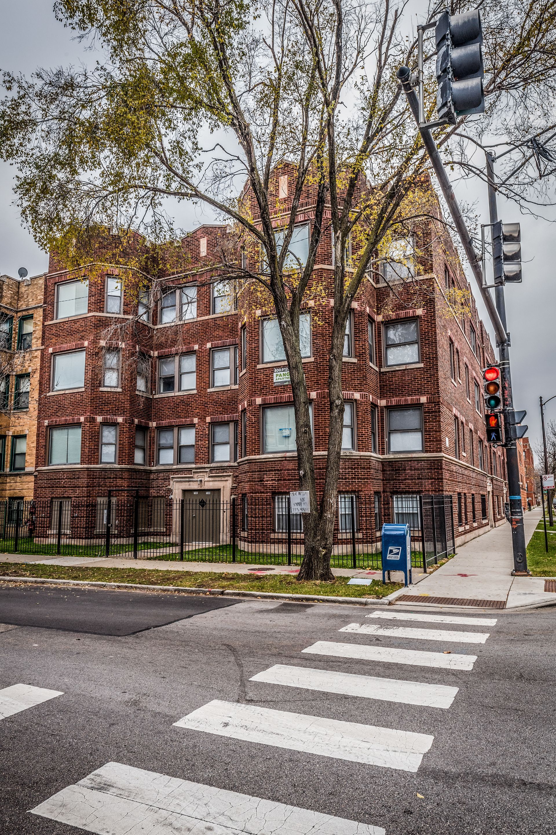 Brick apartment building at a street corner with crosswalk, traffic lights, and a tree. Overcast sky.