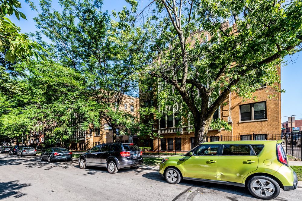 Street view of parked cars in front of apartment buildings on a sunny day. Green trees provide shade.