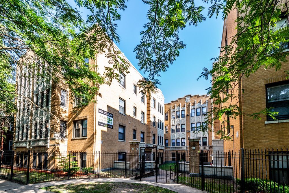 Apartment buildings, beige brick and white accents, surrounded by trees and a black fence, sunny day.