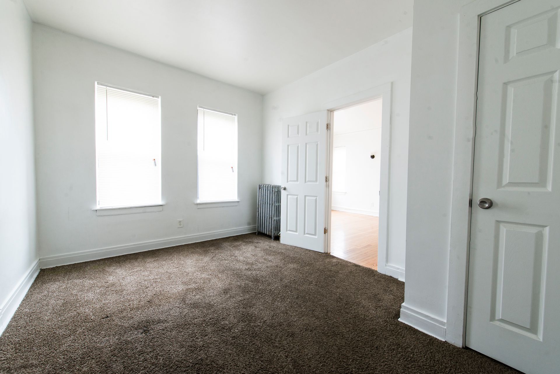 Empty room with brown carpet, white walls, and two windows. A door leads to another room.
