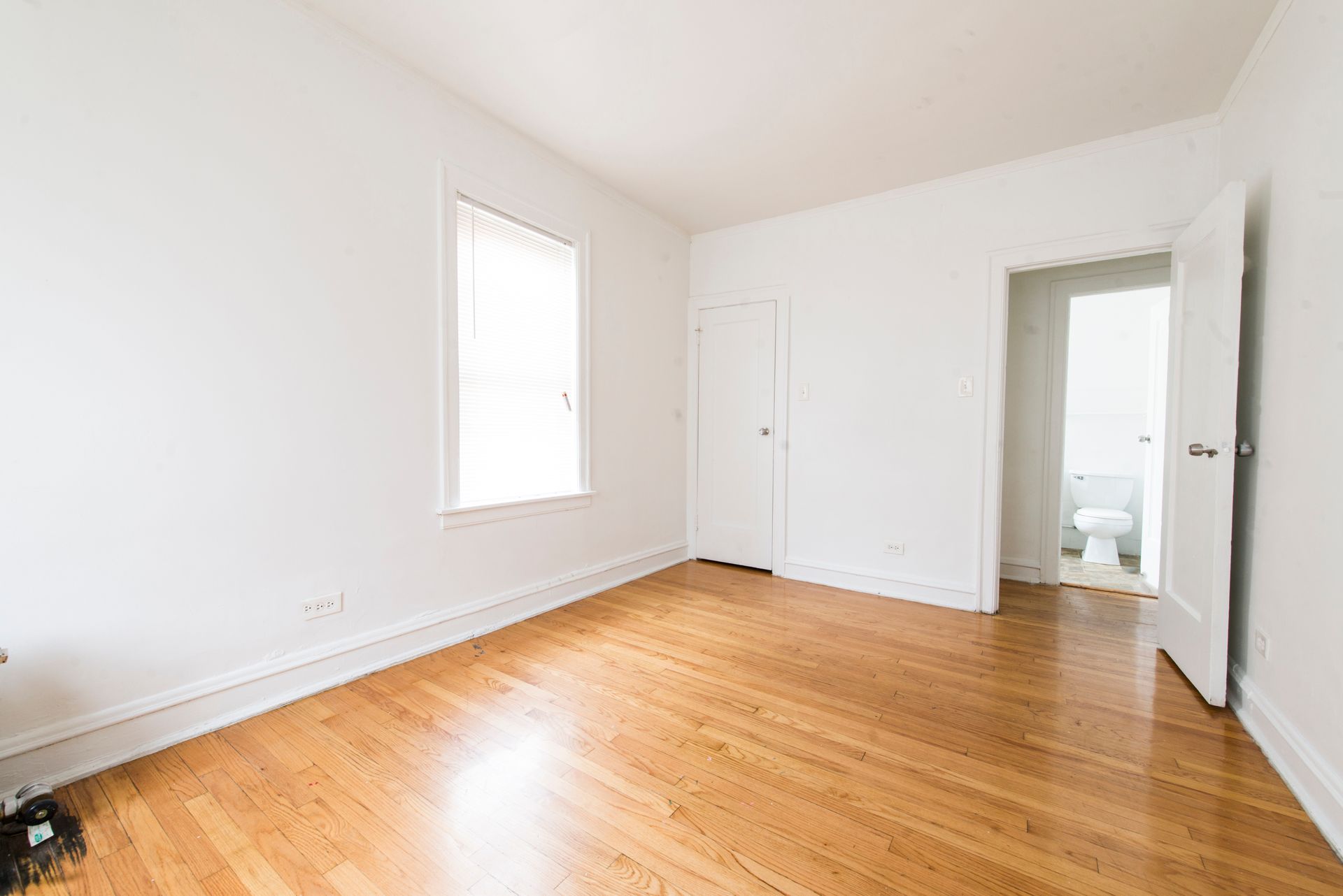 Empty room with wood floors, white walls, closet, window, and open doorway to a bathroom with a toilet.