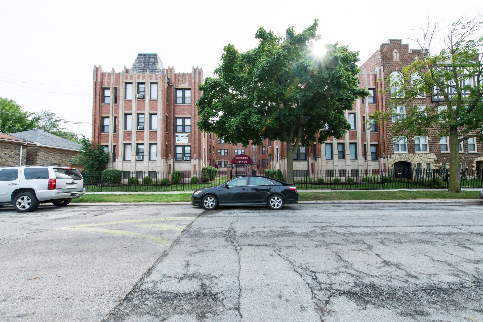 Apartment buildings on a city street, with cars parked in front. Gray sky.