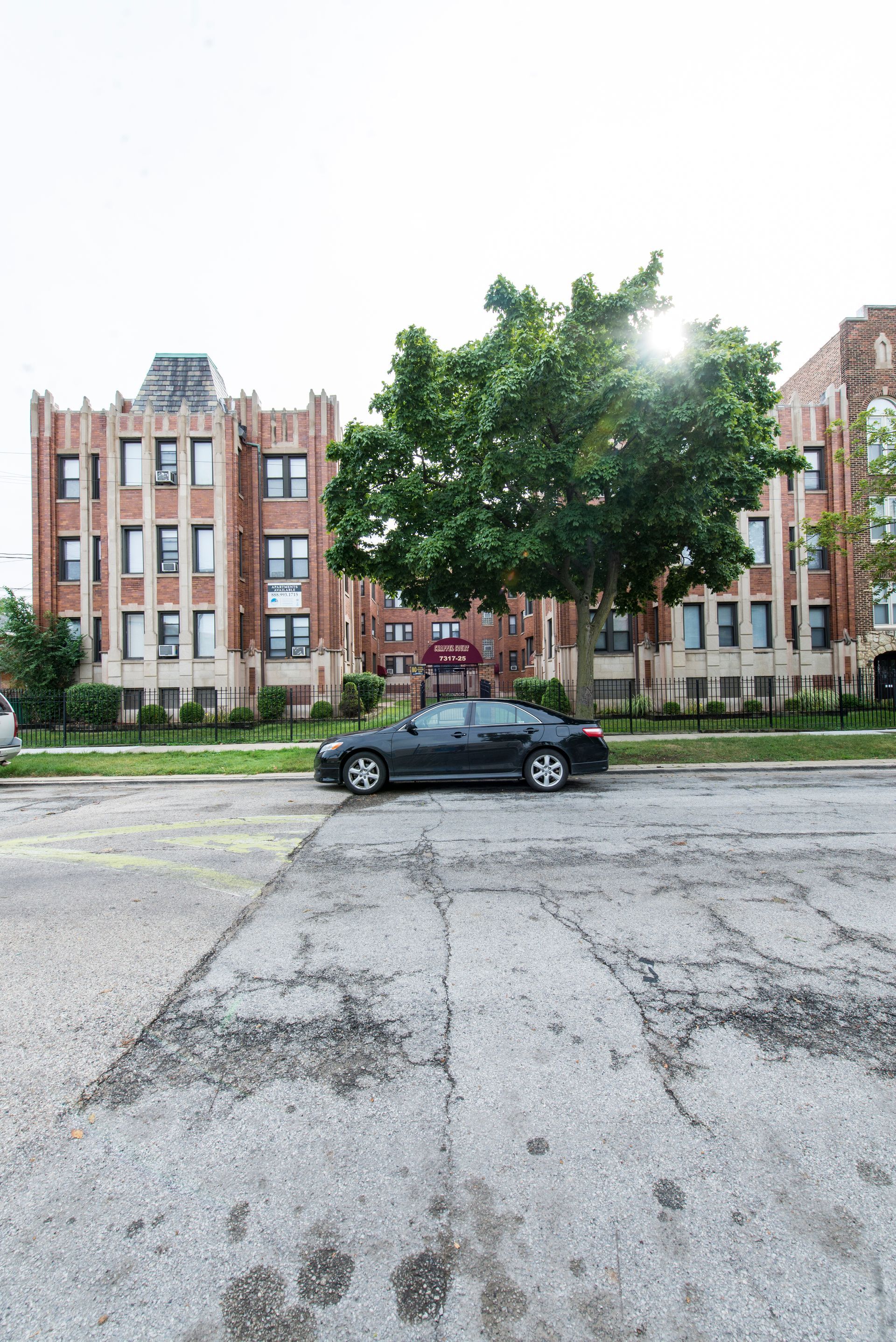 Dark car parked in front of a brick apartment building with a large tree.