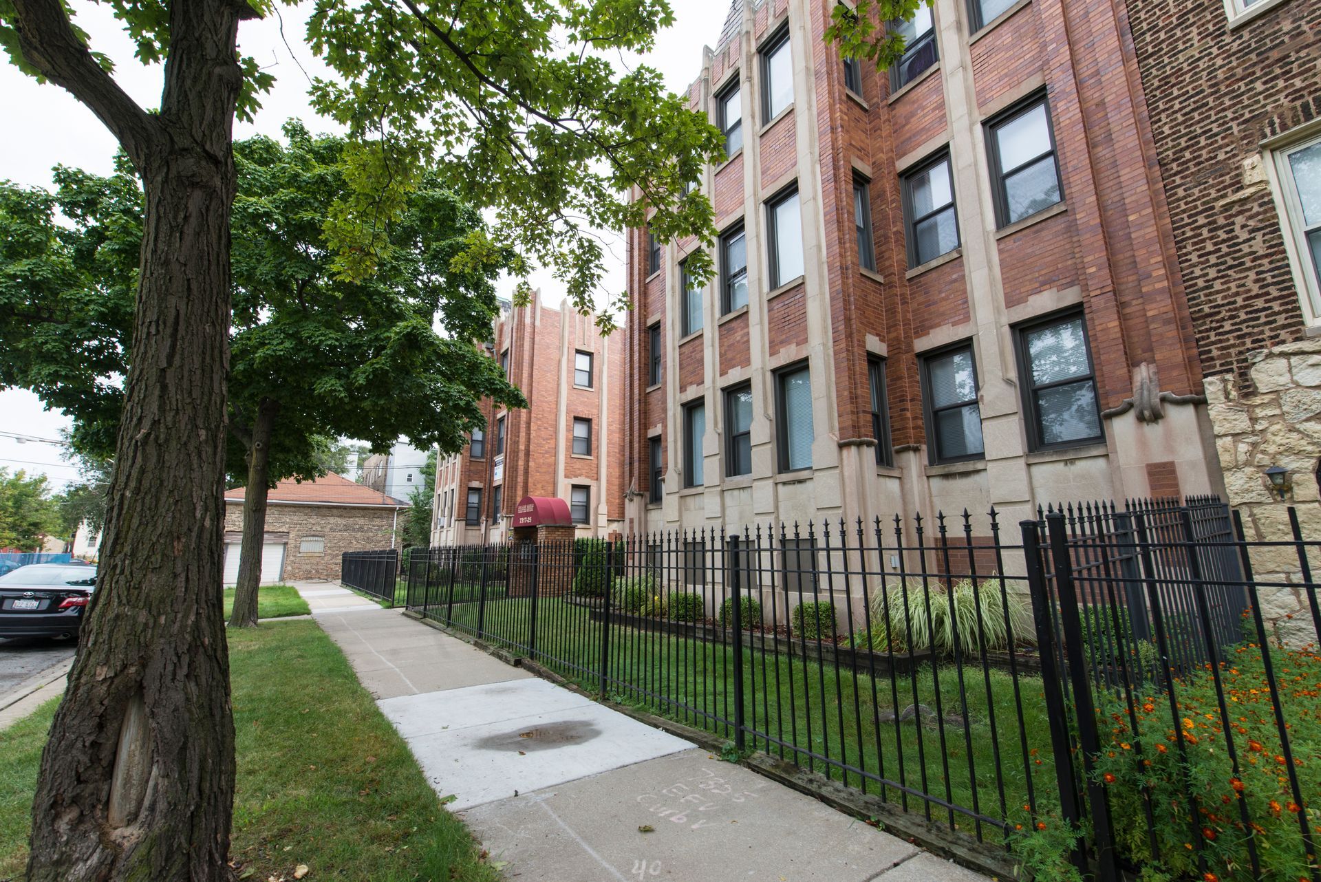 Brick apartment buildings along a sidewalk, with a black fence and trees.