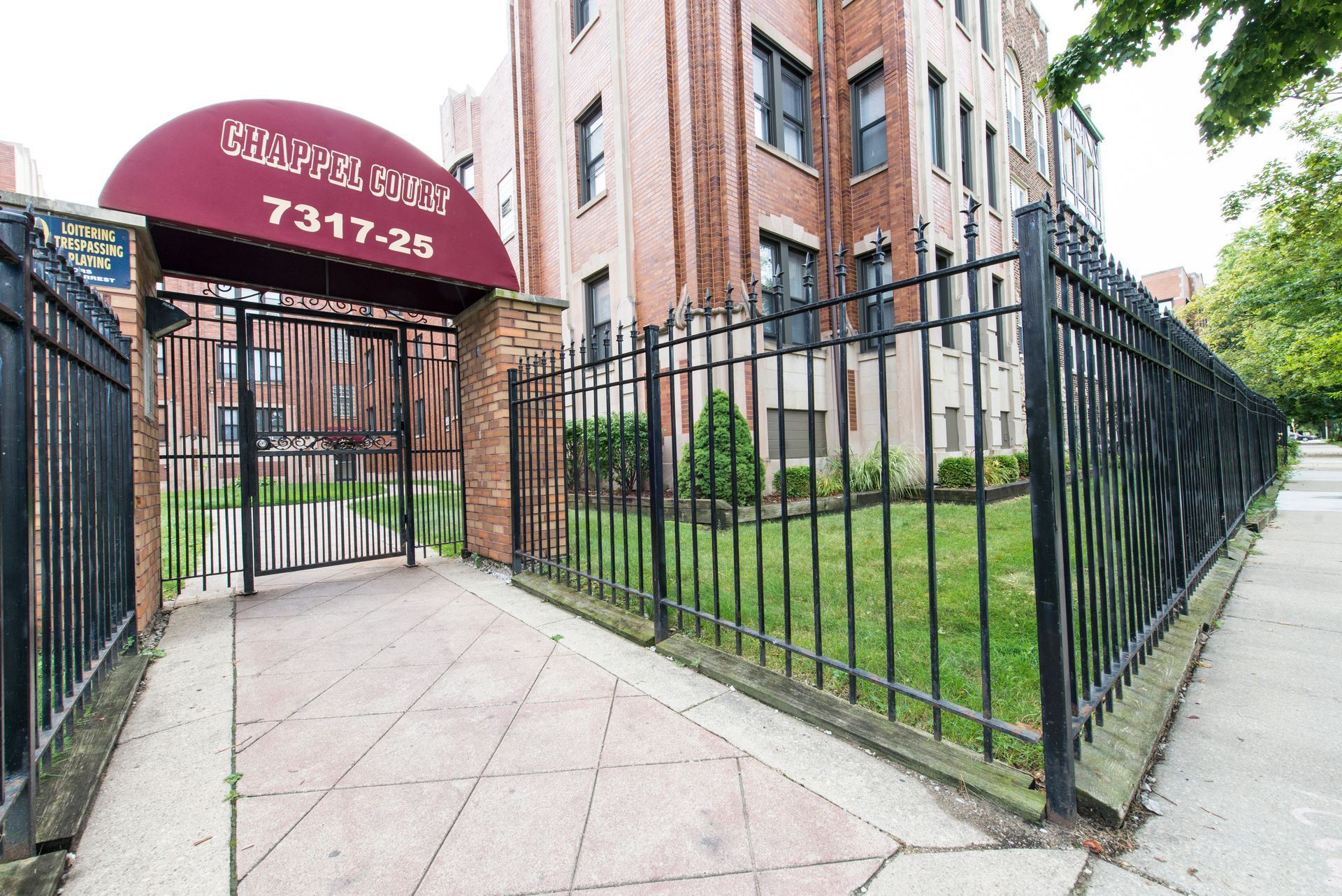 Apartment building entrance with burgundy awning, wrought iron fence, and brick facade.