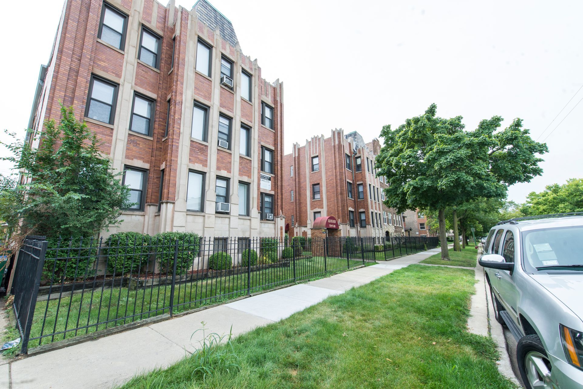Multi-story brick apartment buildings with wrought iron fence and parked car on a grassy street.