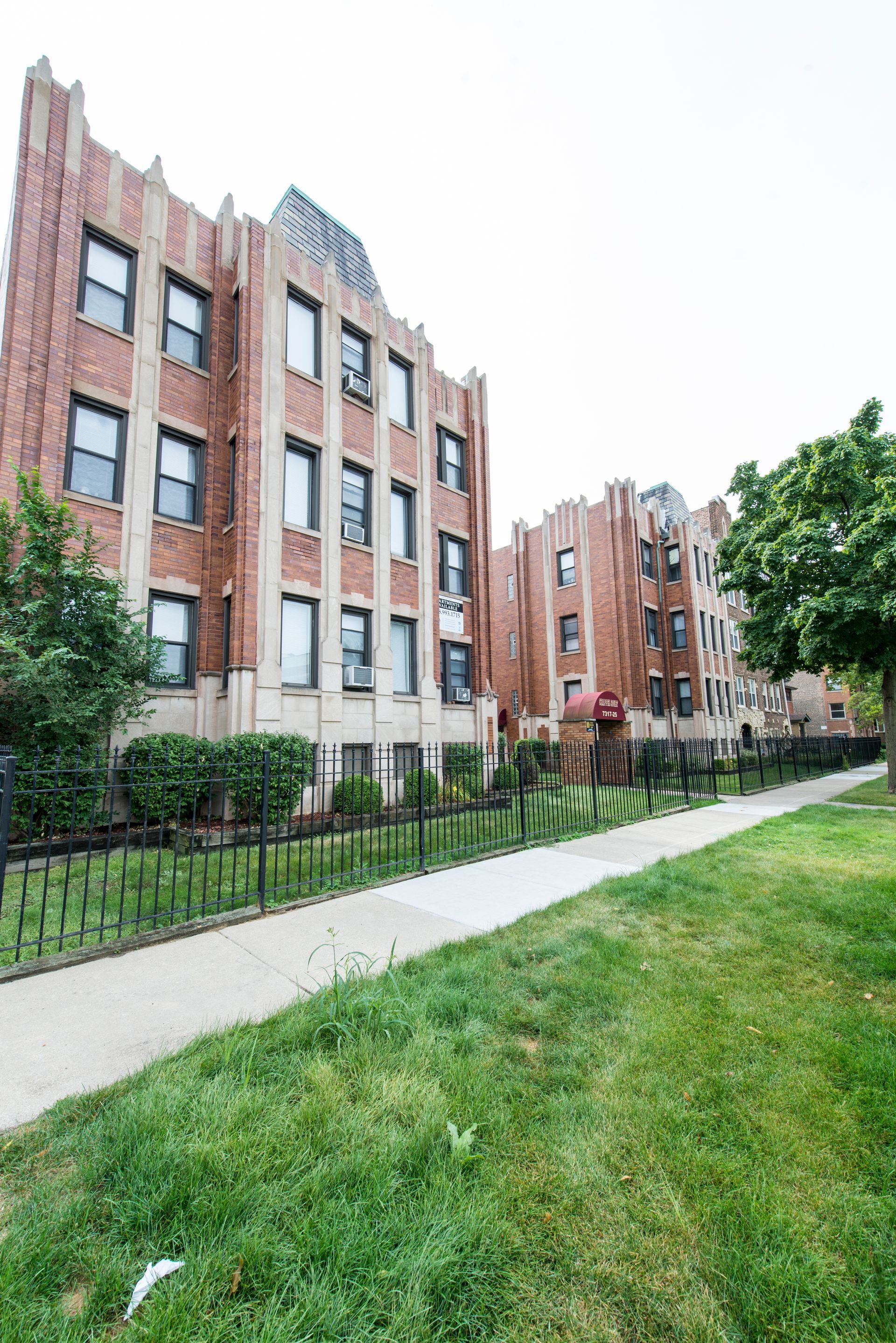 Brick apartment buildings with dark windows sit behind a wrought iron fence and sidewalk on a grassy street.