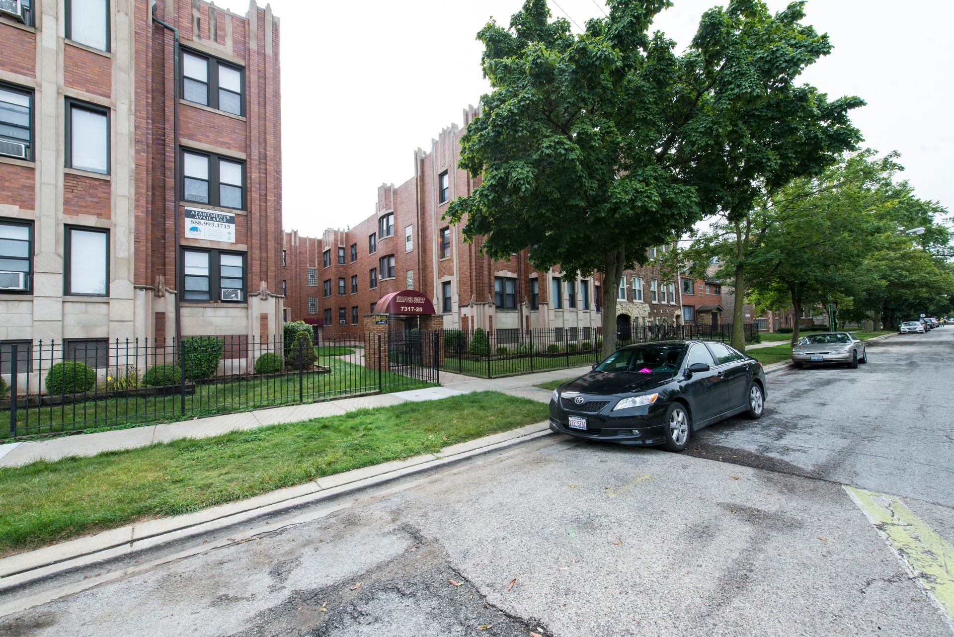 Brick apartment buildings on a city street; a dark car parked on the street; a tree on the right.