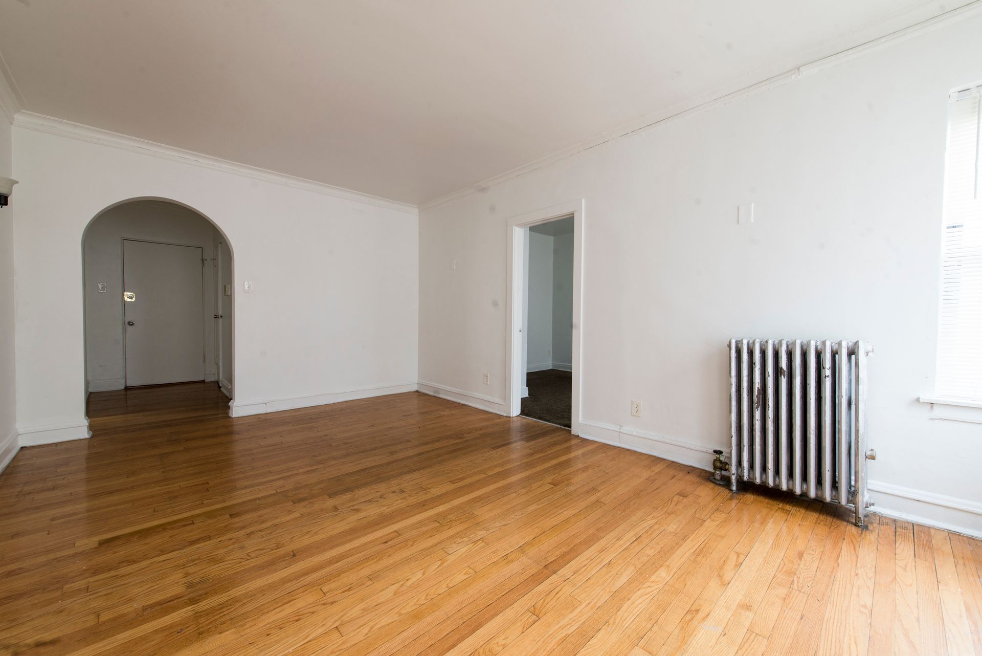 Empty room with wood floors, white walls, arched doorway, radiator, and doorway to another room.