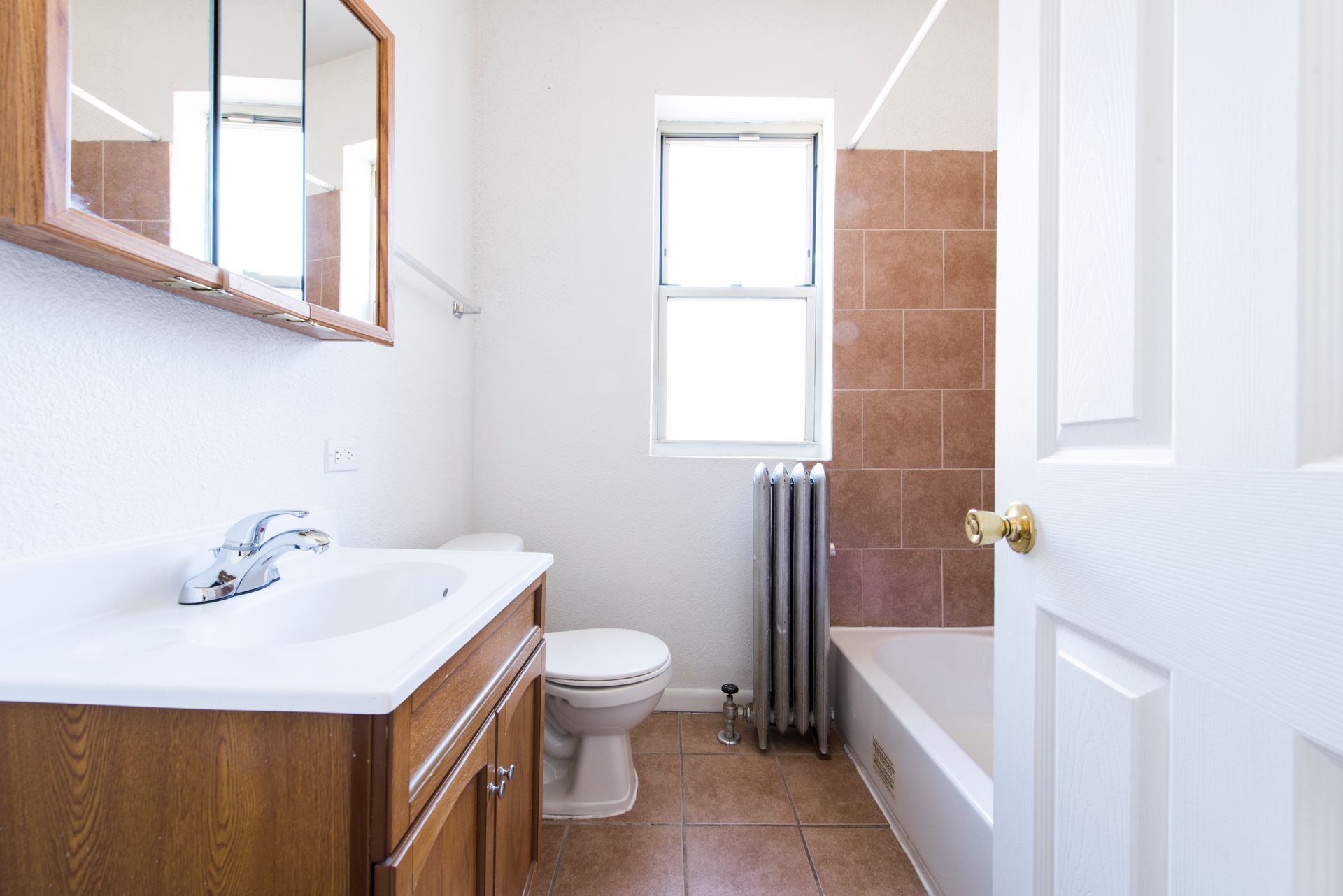 Bathroom with a white sink, toilet, and bathtub. Brown tile and cabinet.  A window is open.  The door is open.