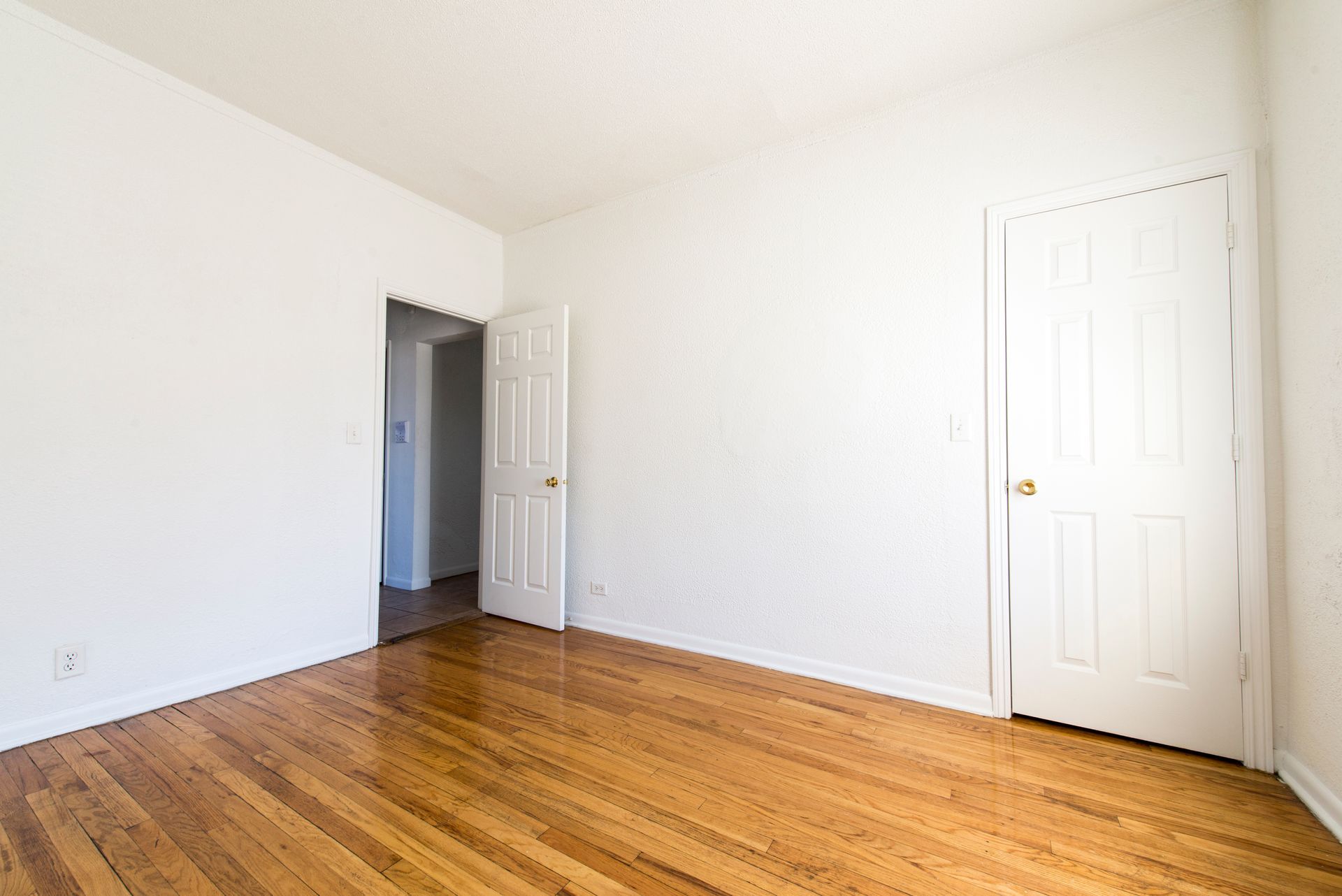 Empty room with hardwood floors, white walls, two white doors, one open to a hallway.