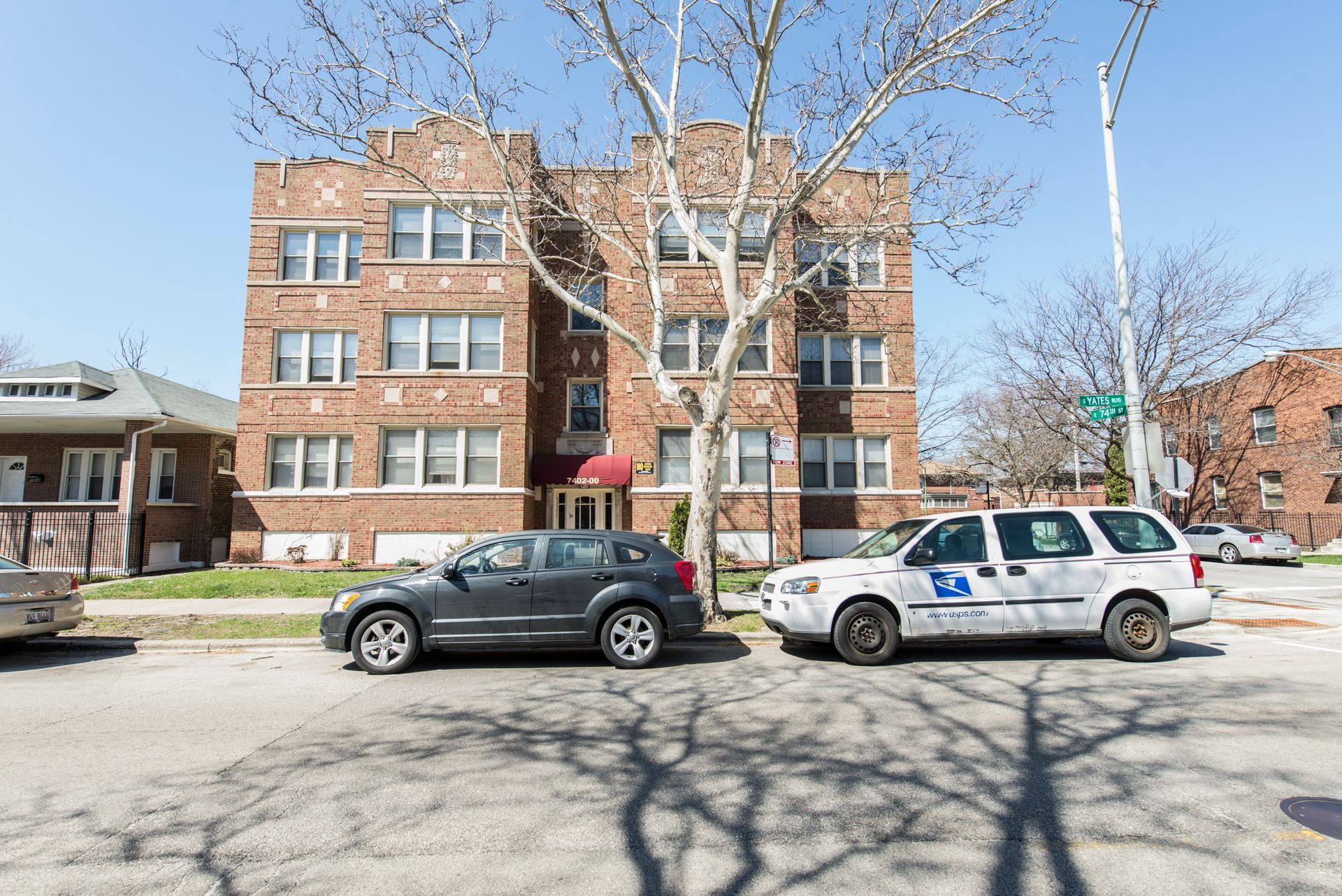 Brick apartment building with parked cars on a sunny street, tree in front.