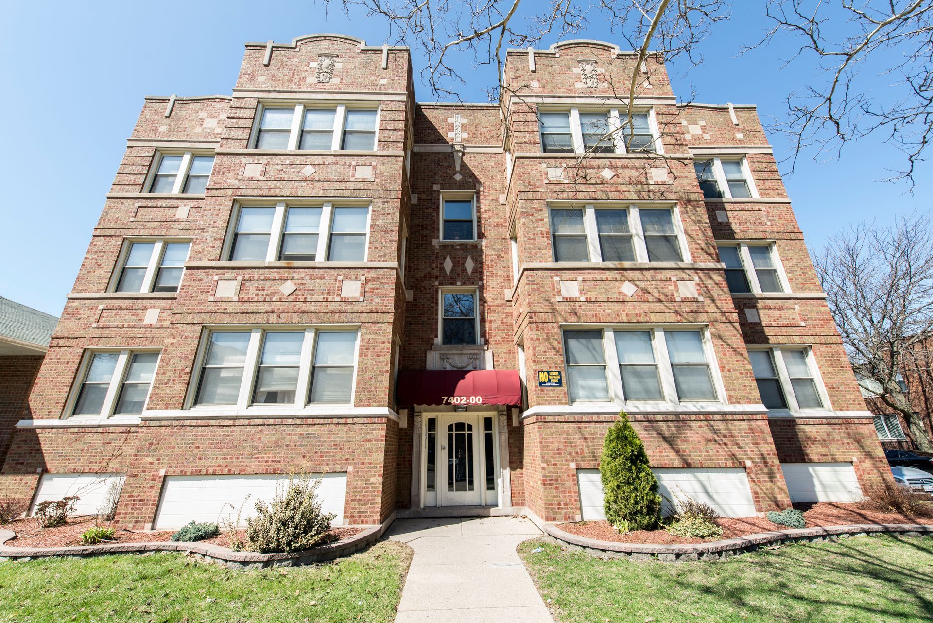 Brick apartment building with white-framed windows, red awning over entrance, and small yard with greenery.