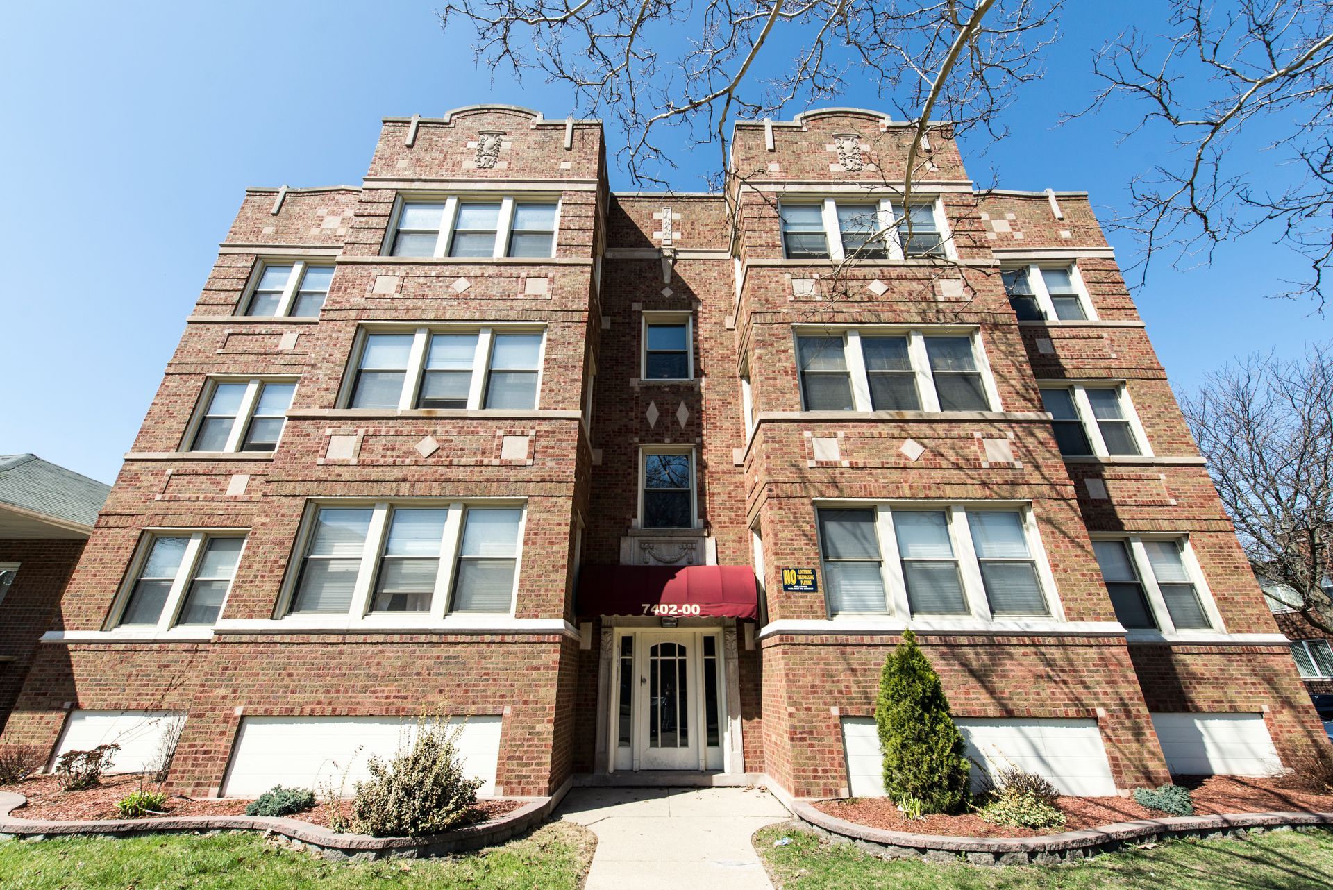 Brick apartment building with white-framed windows, red awning, and front door, under a blue sky.
