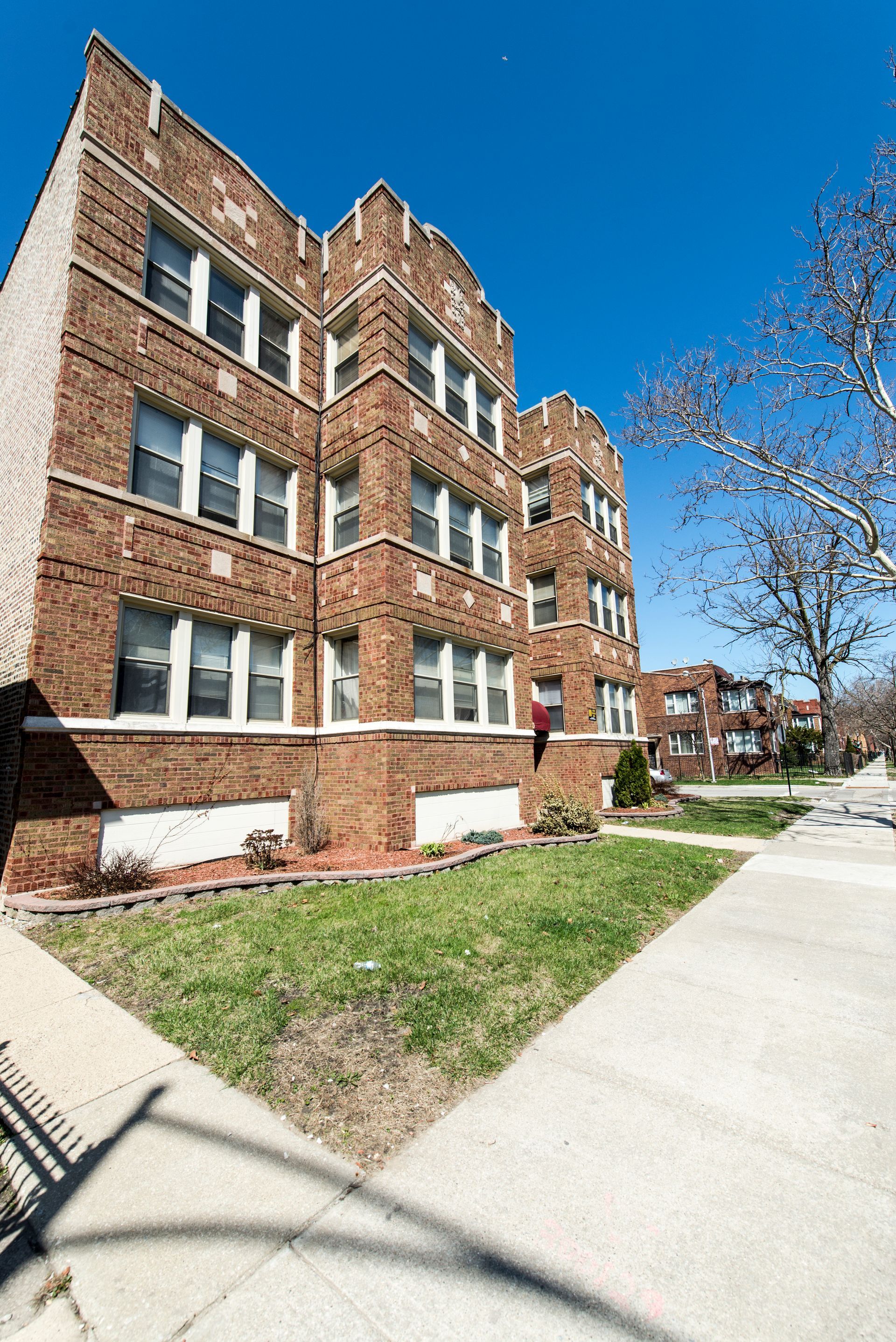 Brick apartment building, three stories, with white-framed windows, against a blue sky, on a sunny day.