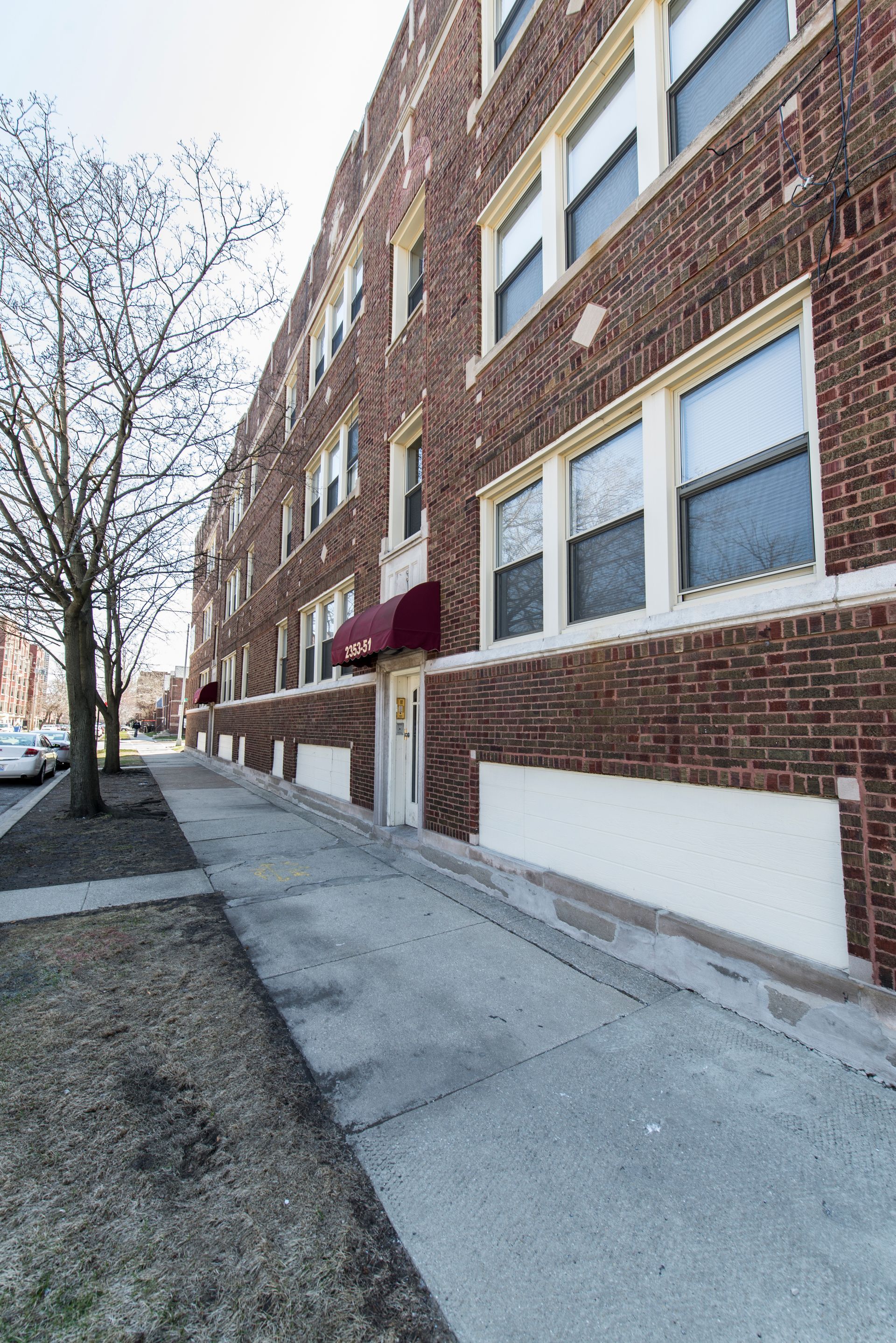 Brick apartment building with ramp to the entrance, sidewalk, and tree on a sunny day.