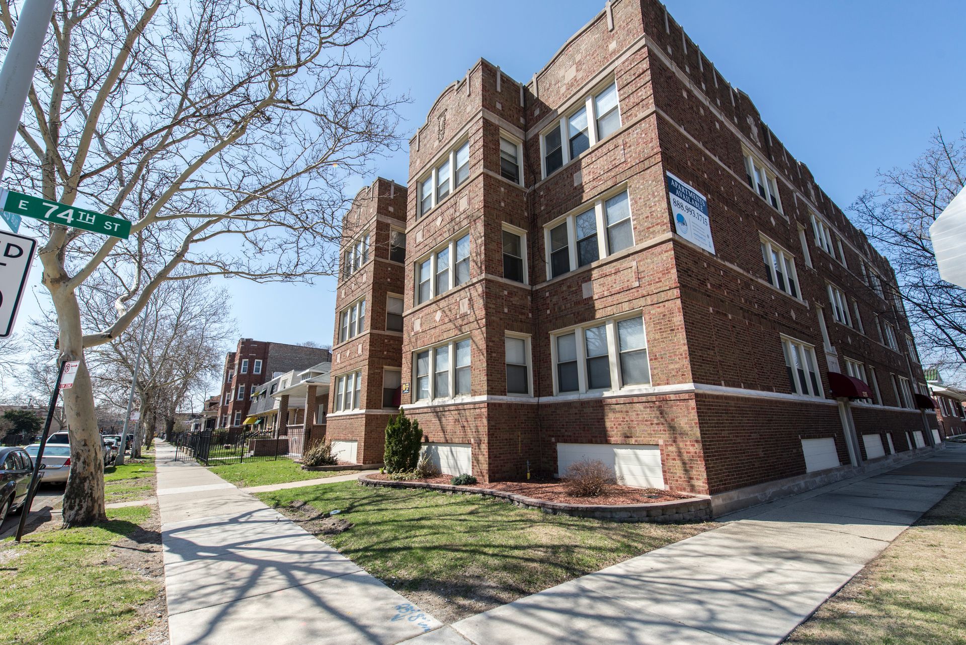 Brick apartment building on a sunny street corner with a green street sign and trees.