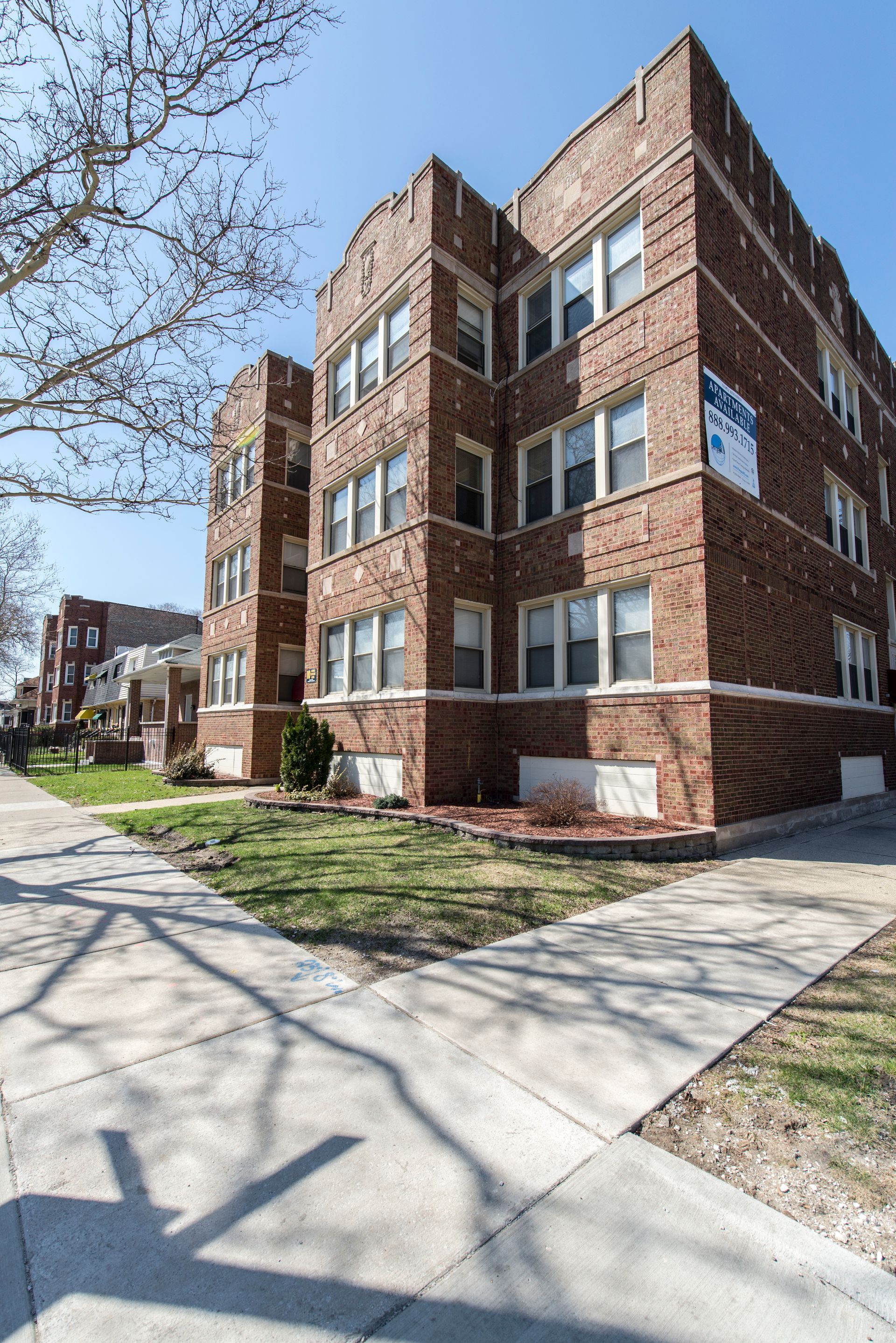 Brick apartment building on a sidewalk with sparse green grass and bare tree branches.