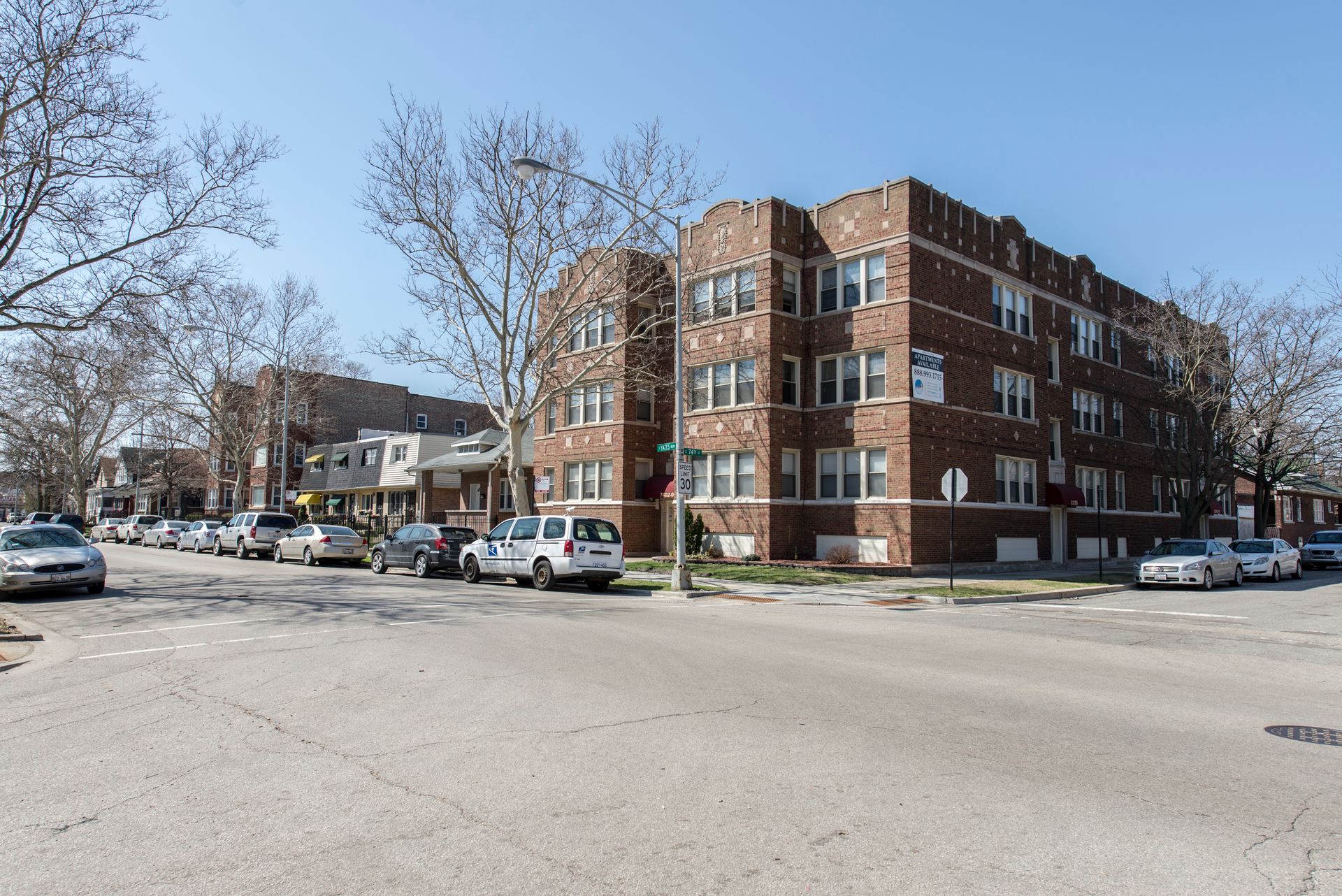 Brick apartment buildings on a city street with parked cars and bare trees under a blue sky.