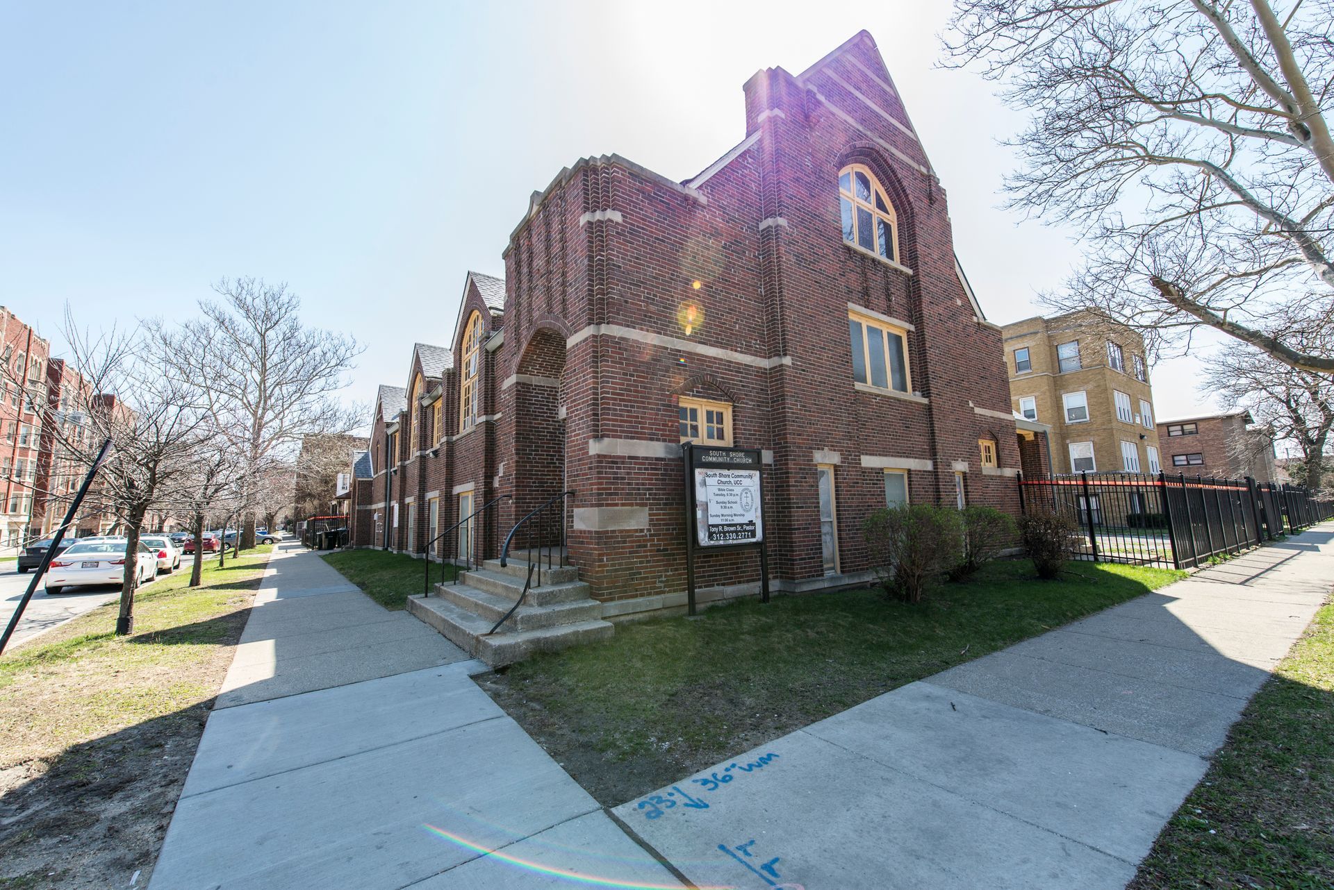 Brick building with arched window and sign, next to a sidewalk and street with parked cars.