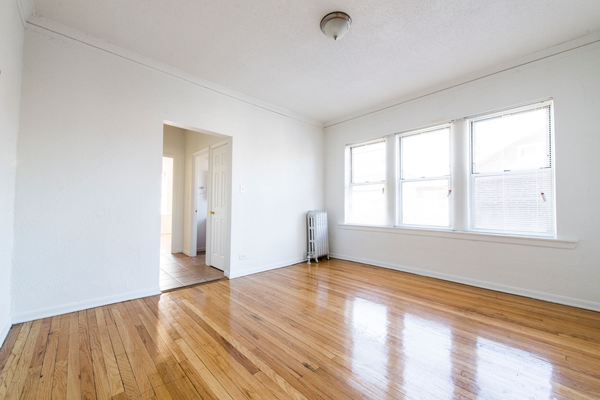 Empty room with hardwood floors, white walls, and three windows. Doorway to another room on the left.