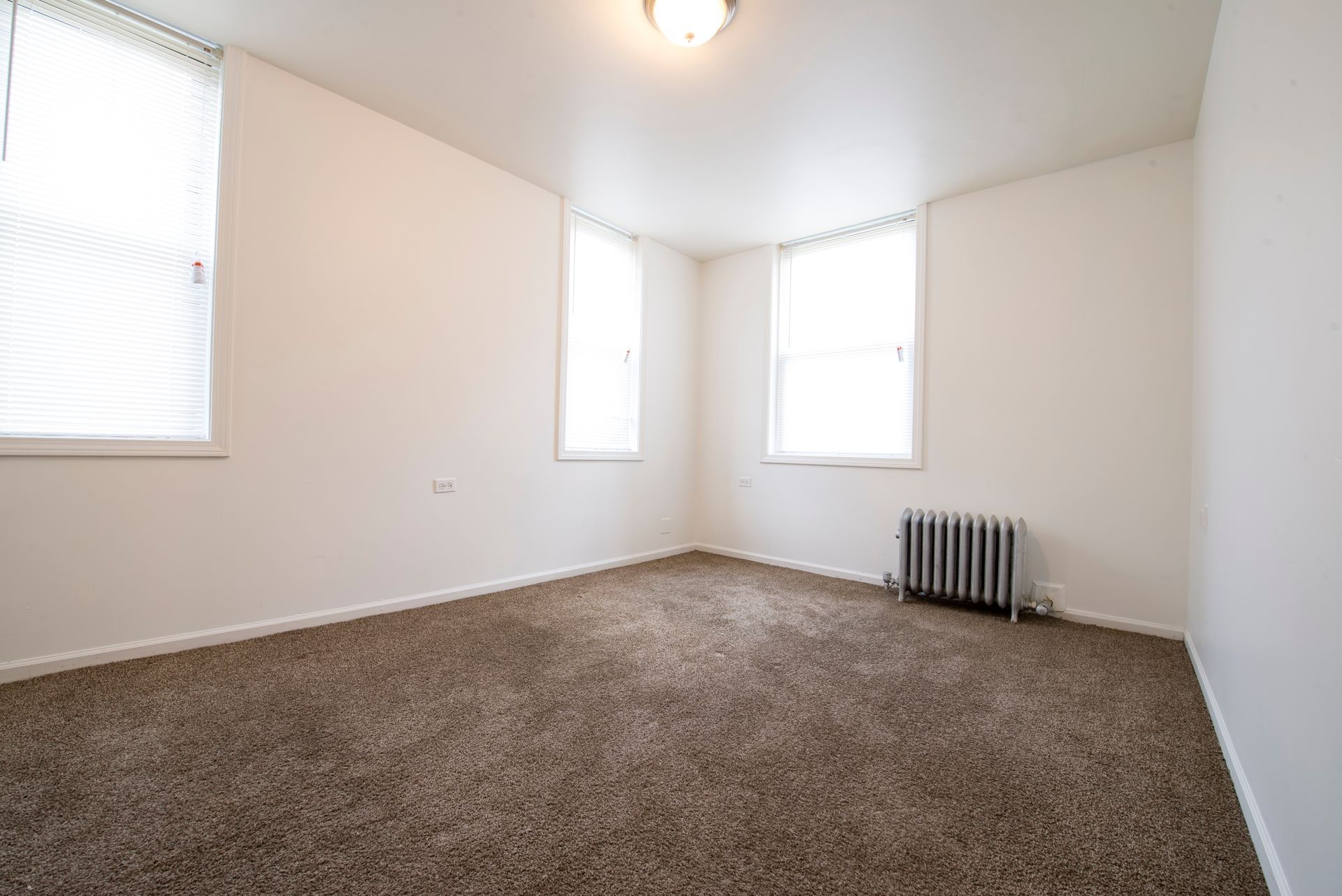 Empty room with brown carpet, white walls, three windows, and radiator.
