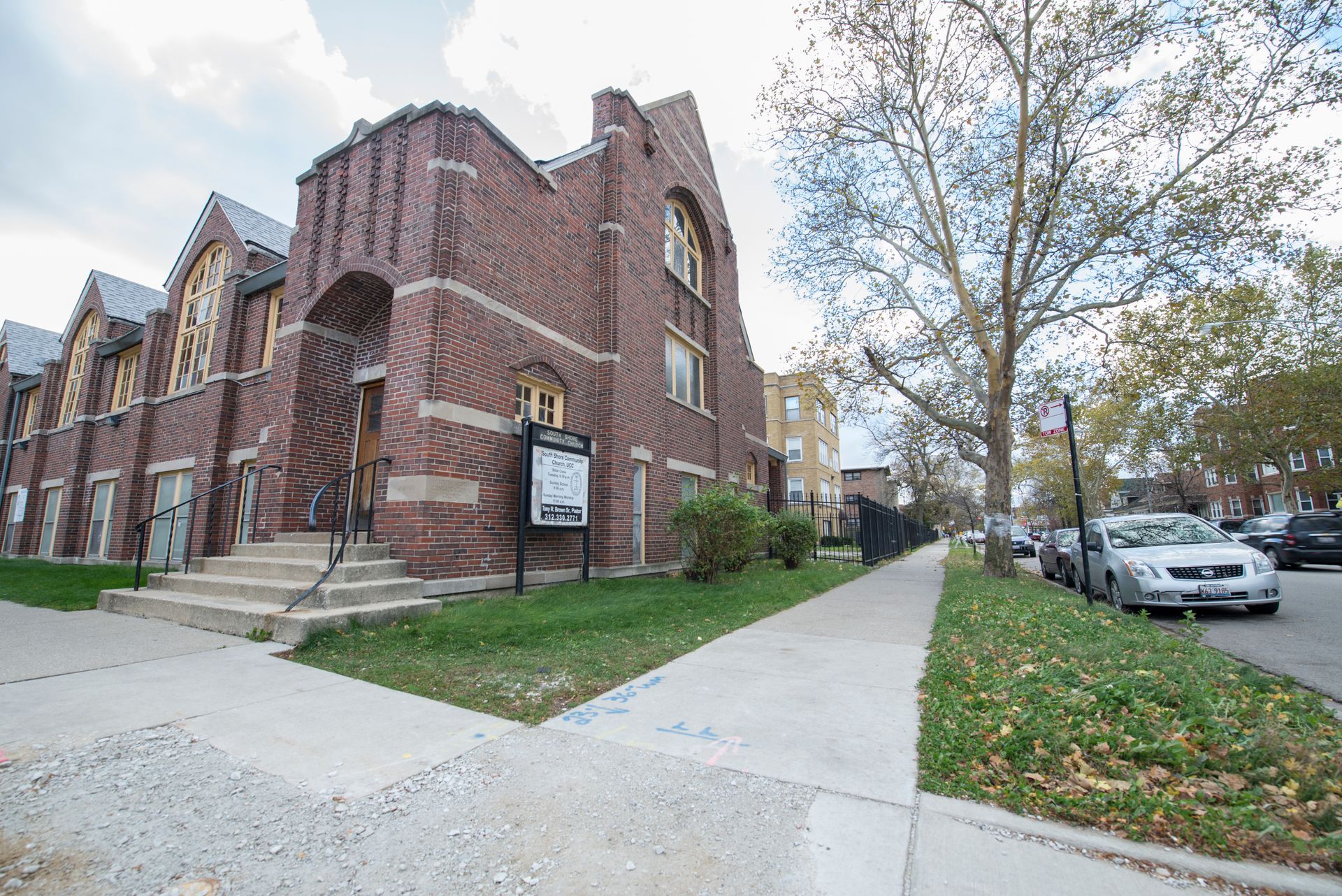 Brick church building with a sidewalk, parked cars, and a tree-lined street.