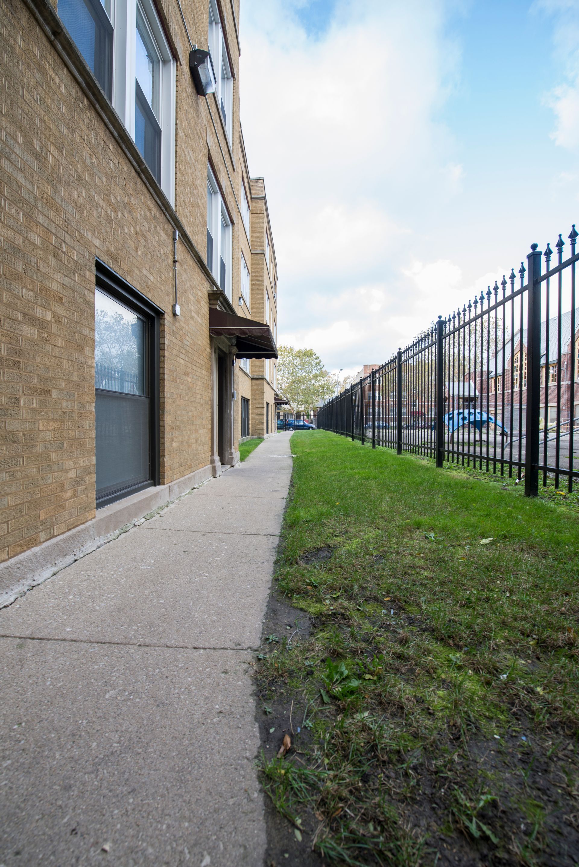 Sidewalk beside a brick building with windows, a grassy area, and a black metal fence on a sunny day.