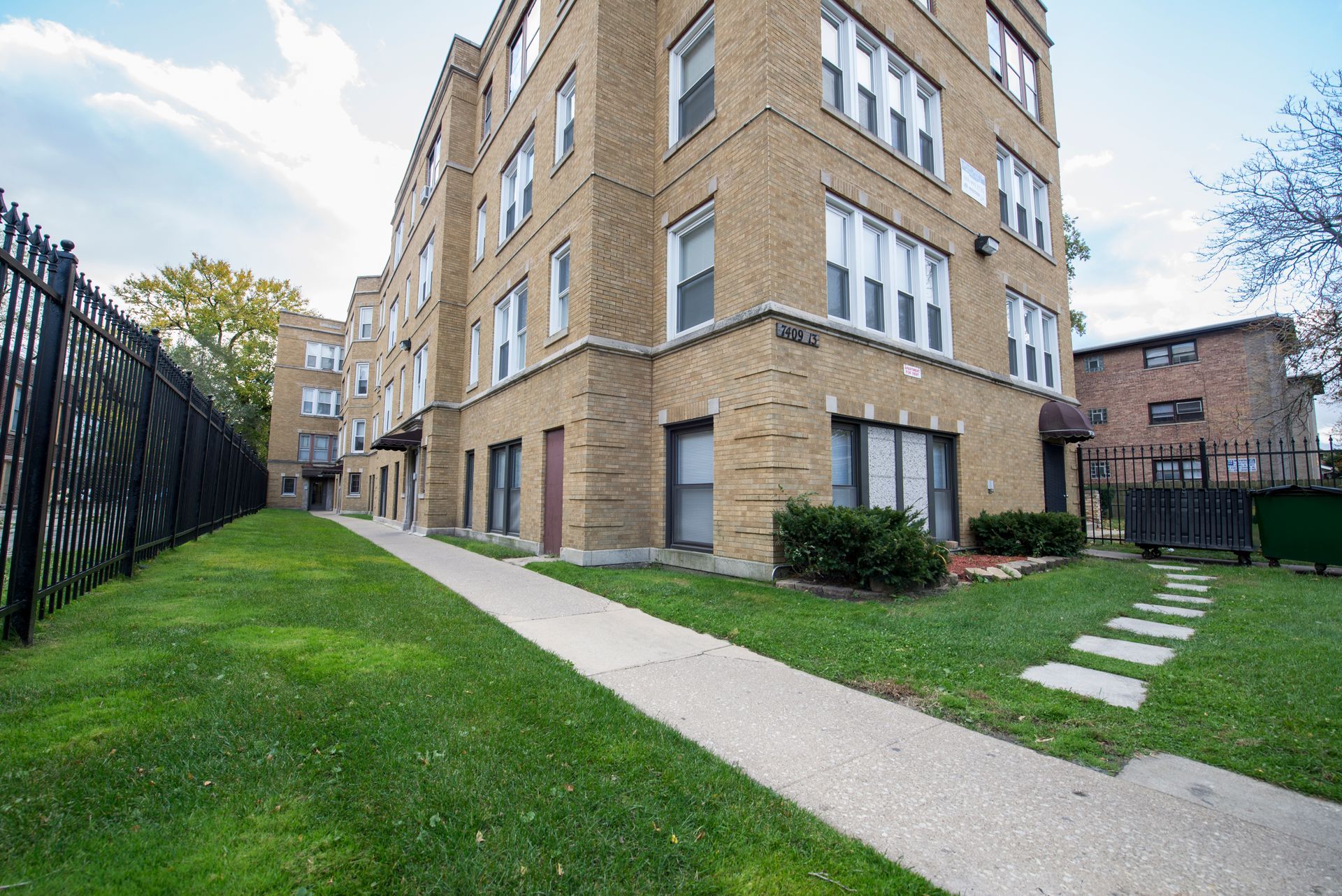 Apartment building with brick exterior, green lawn, and a sidewalk. Black fence runs along the side.