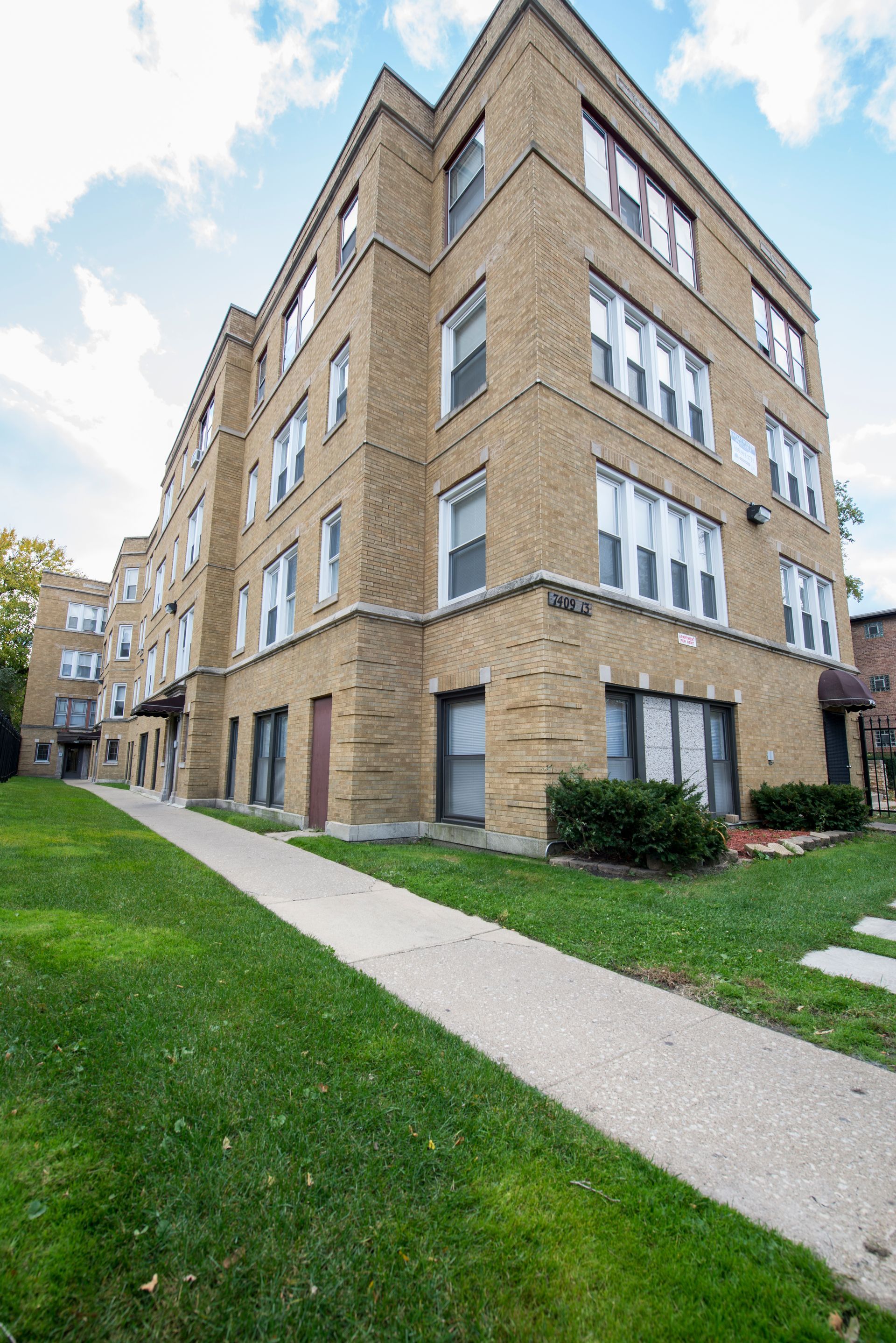 Apartment building with brick exterior, sidewalk, and green lawn on a sunny day.