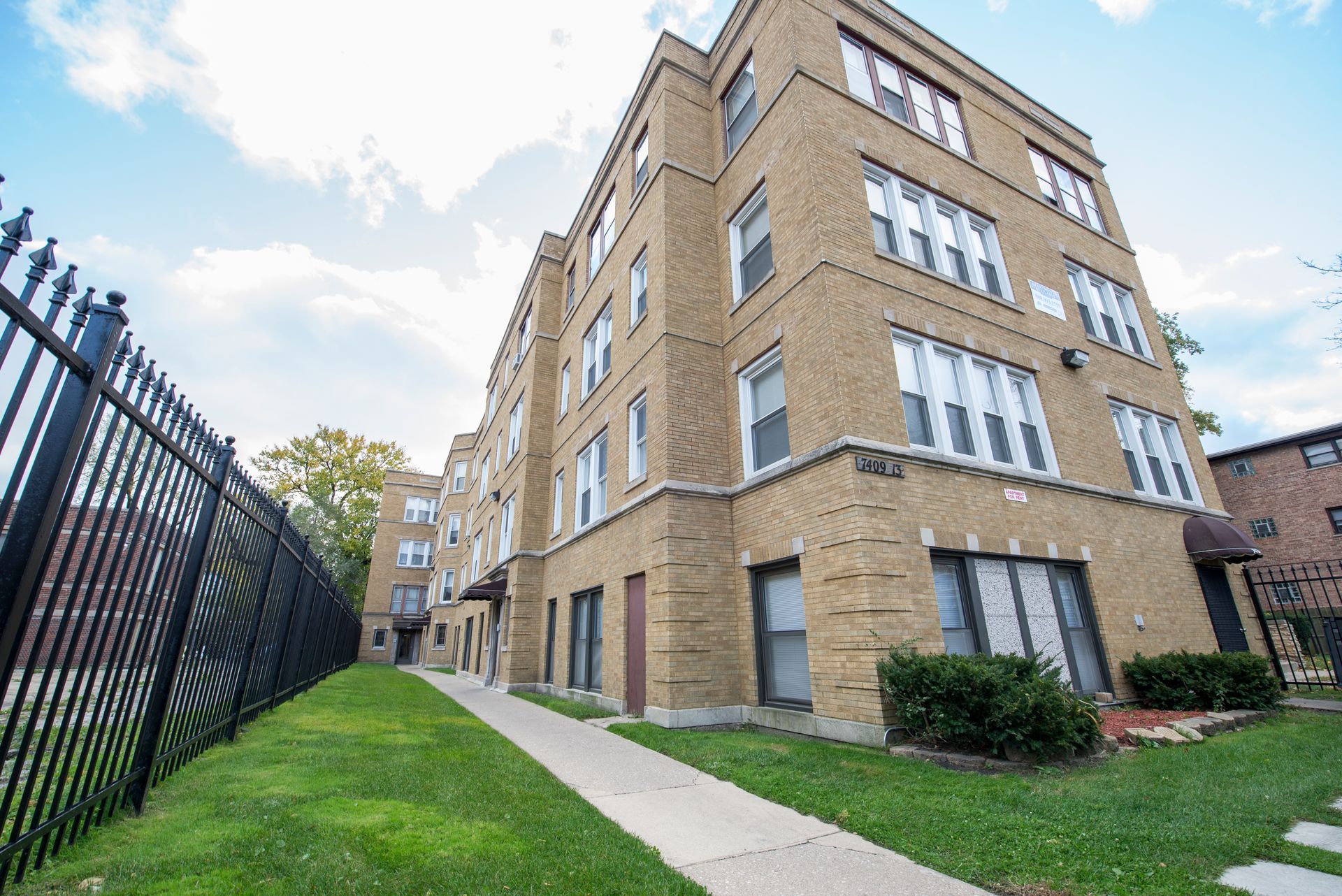 Brick apartment building with black fence, walkway, and blue sky.