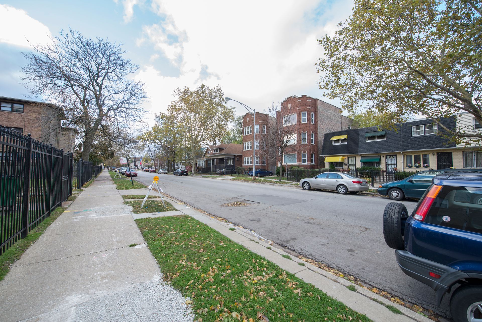 Street view of residential area with houses, cars parked, and sidewalk. Green grass and trees visible.