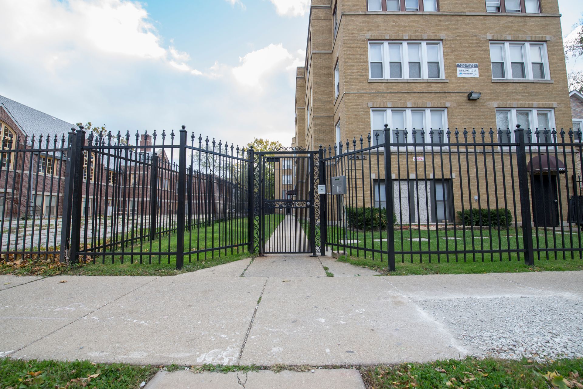 Black metal fence with gate leading to an apartment building. Pavement and grass in foreground. Cloudy sky.