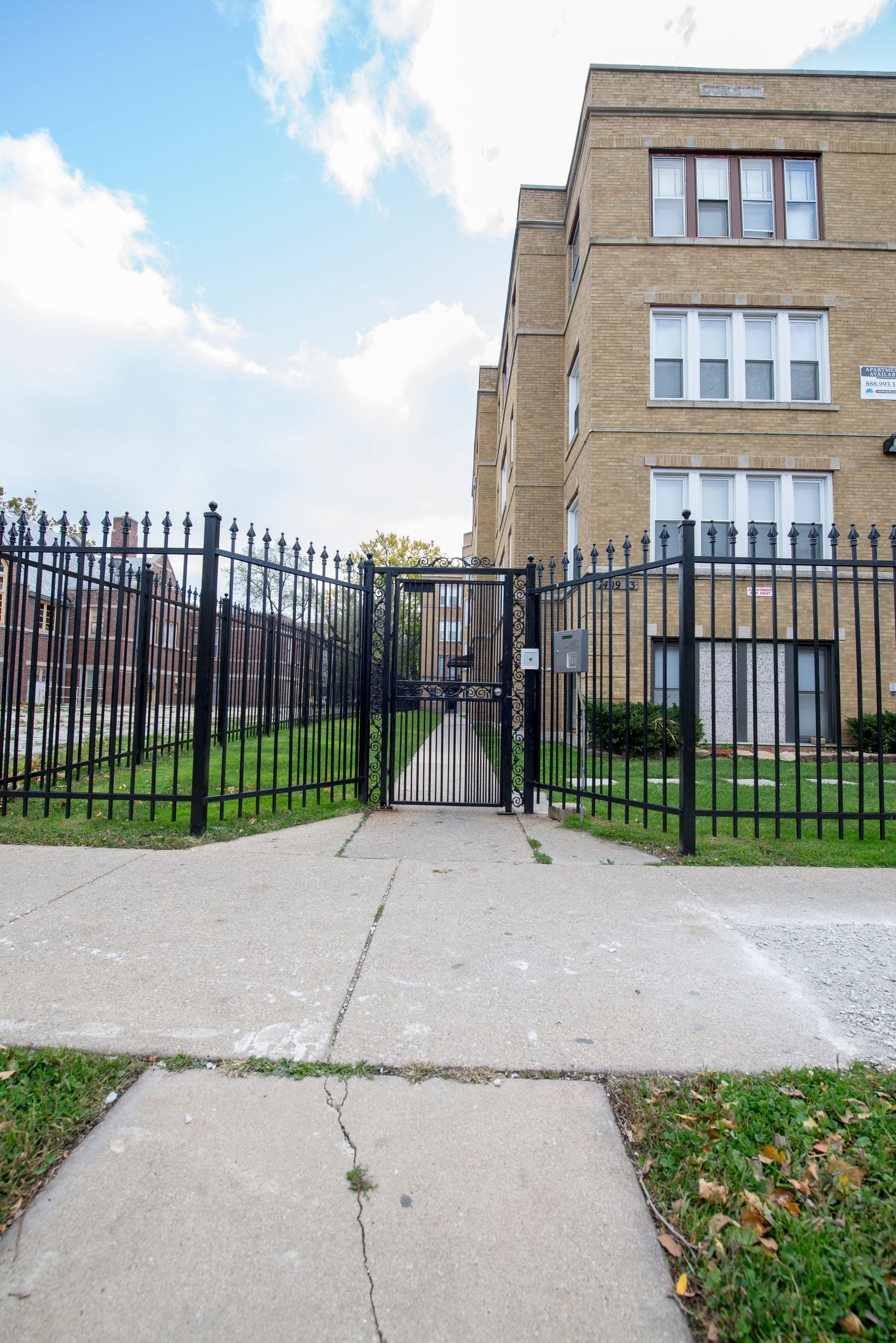 Black metal fence and gate leading to a tan brick apartment building under a blue sky.