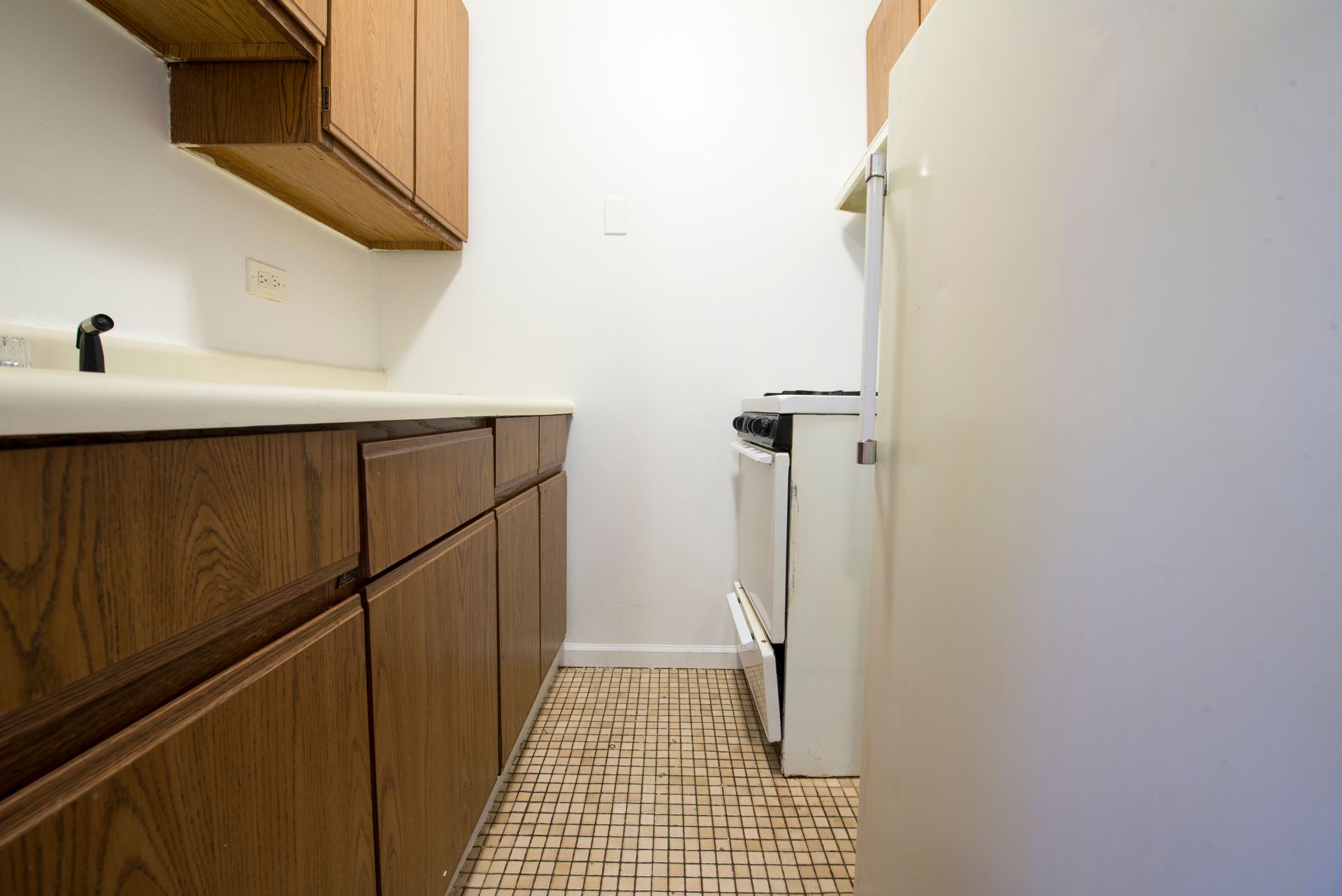 A narrow kitchen with wood cabinets, white appliances, and patterned floor.