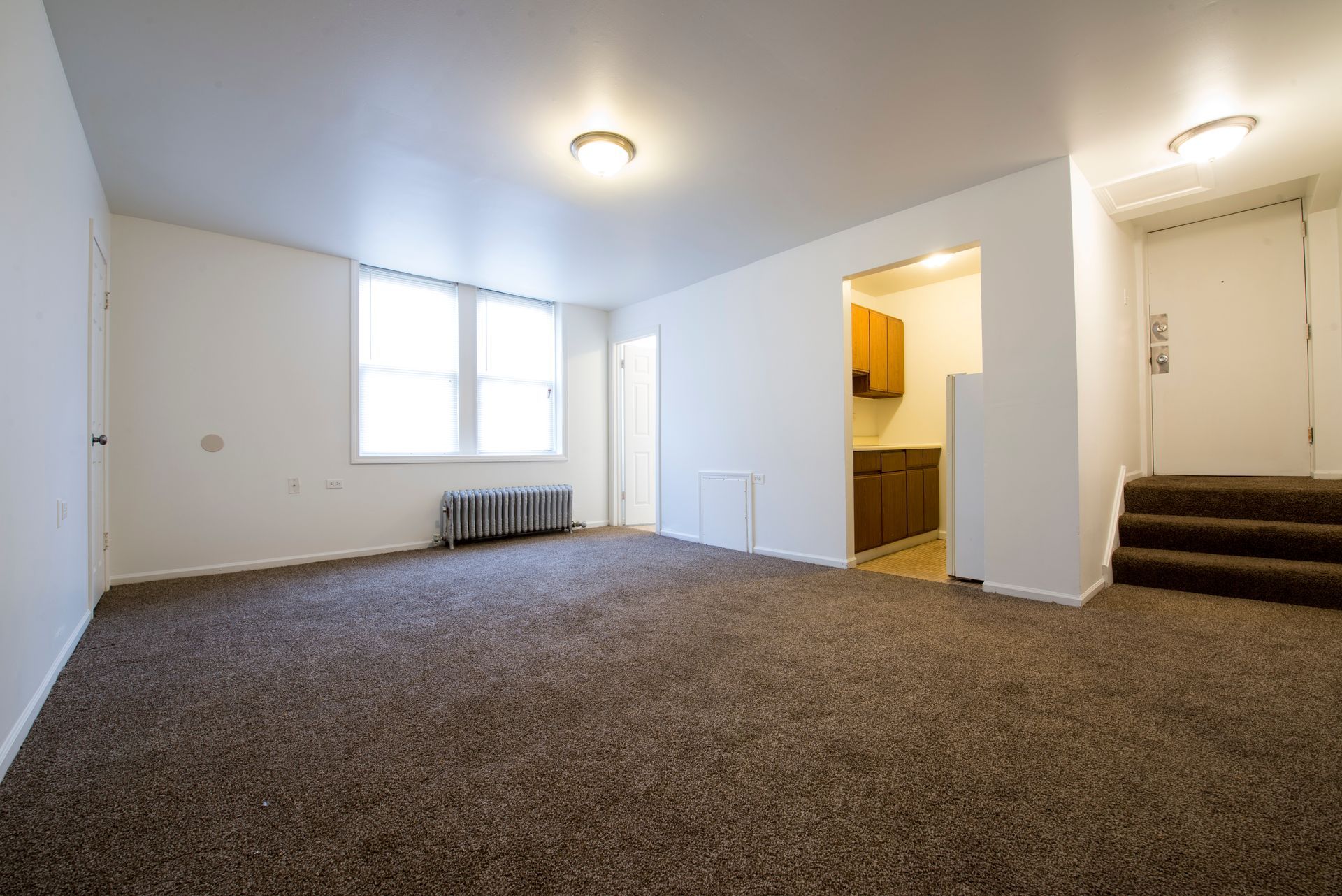 Empty living room with brown carpet, white walls, and a small kitchen visible.
