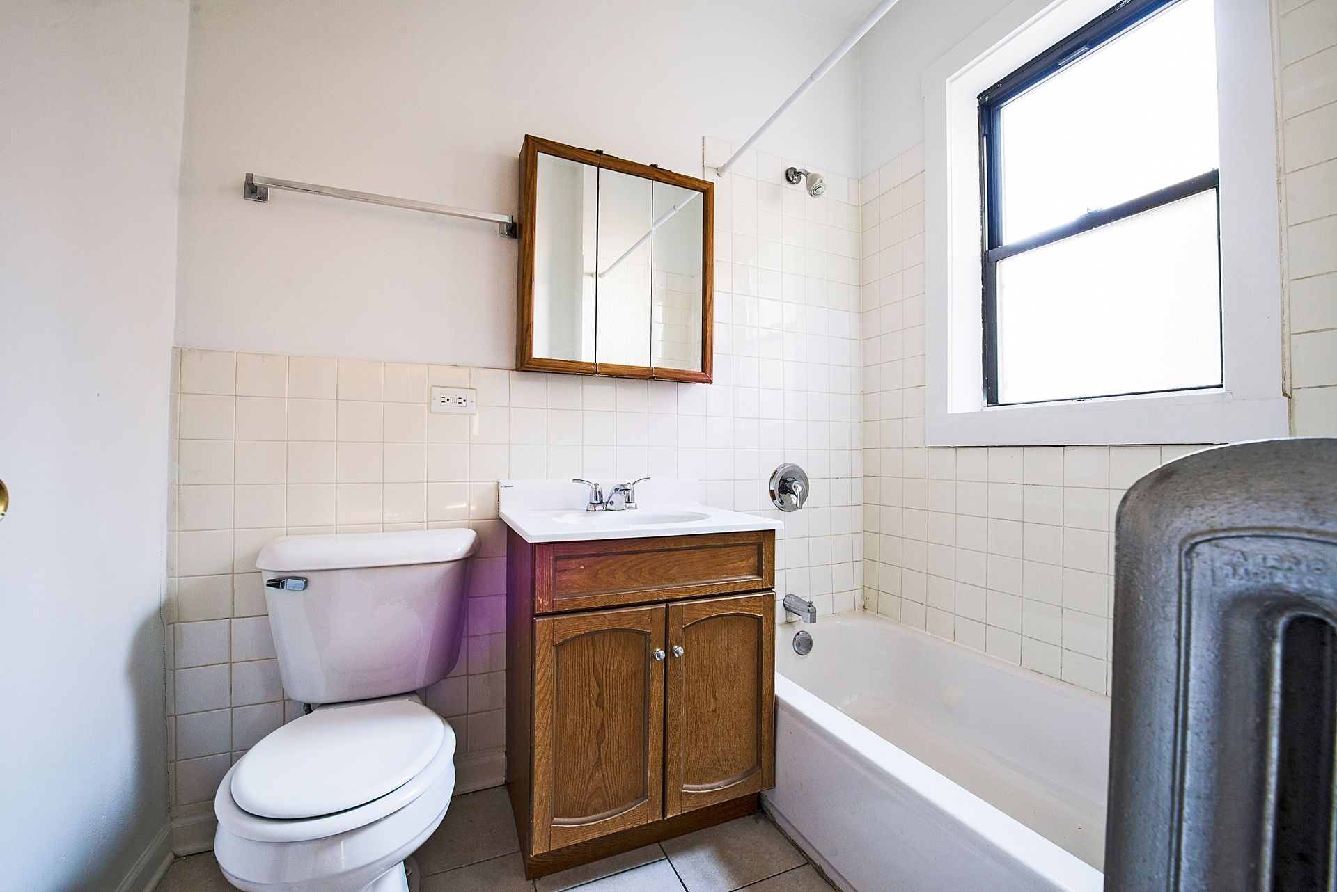 Small, white-tiled bathroom with a toilet, sink, mirror, and bathtub. A window is on the right.