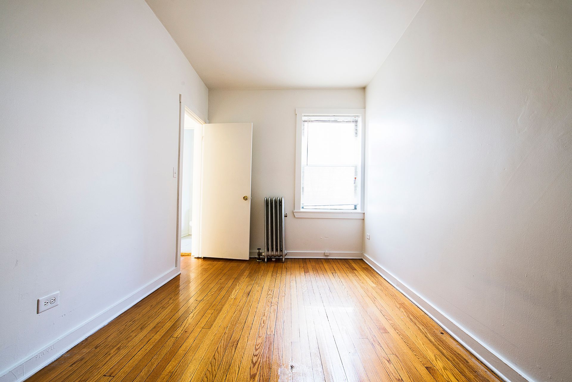 Empty room with wood floor, white walls, closed door, and window.