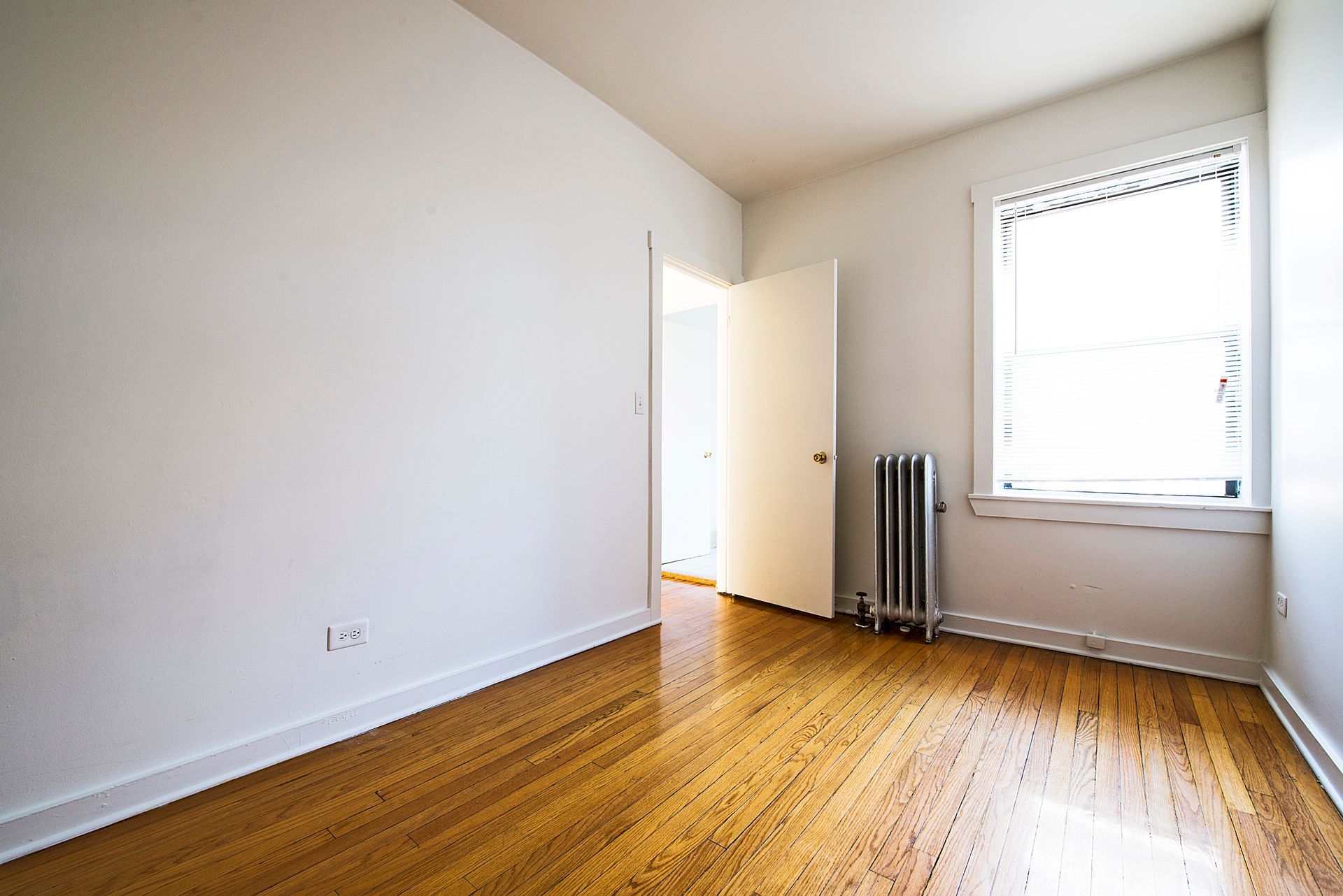 Empty room with wood floor, white walls, open door, radiator, and window.